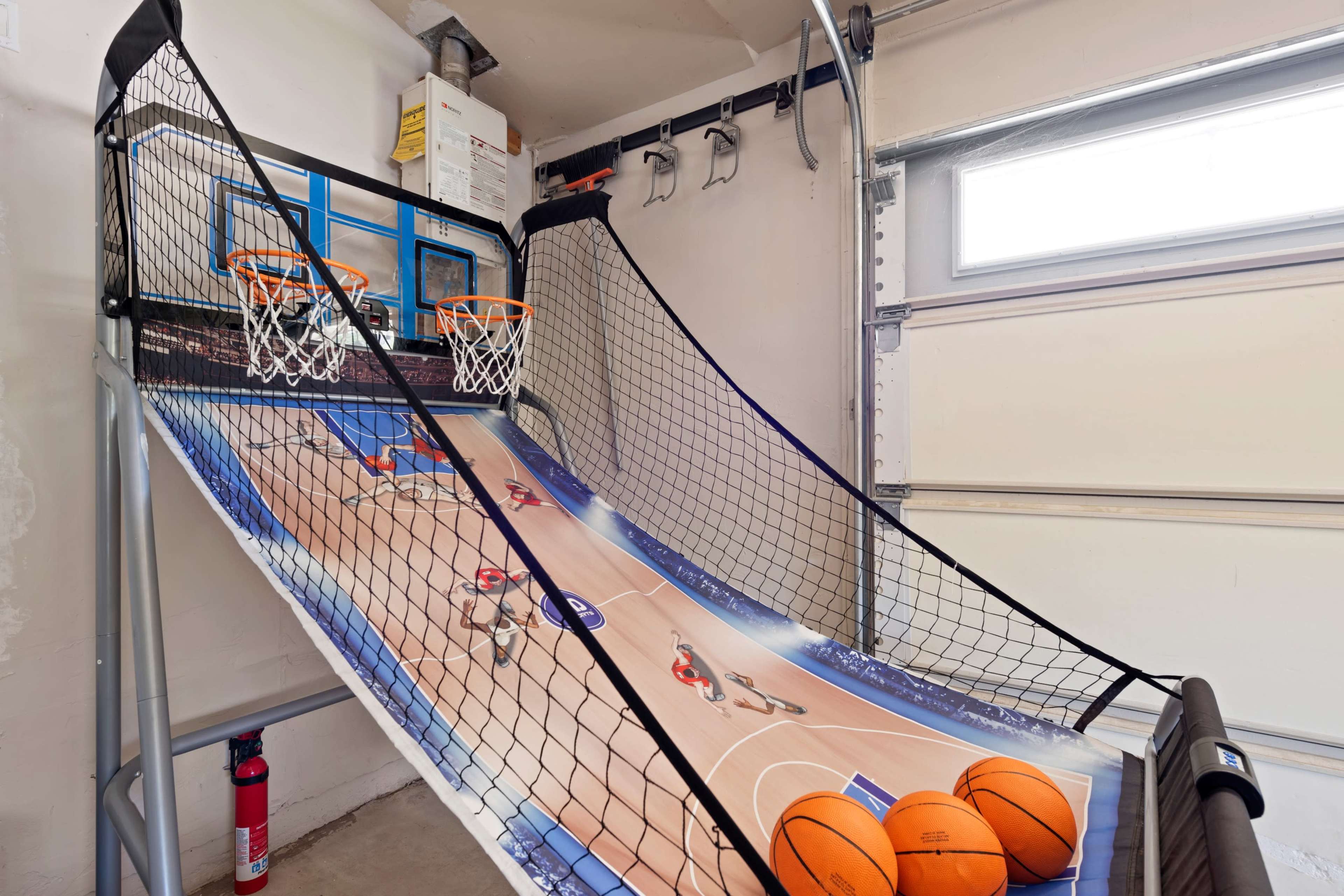 The image shows a home basketball arcade game set up in a garage with two basketball hoops, a sloped playing surface, and several orange basketballs.