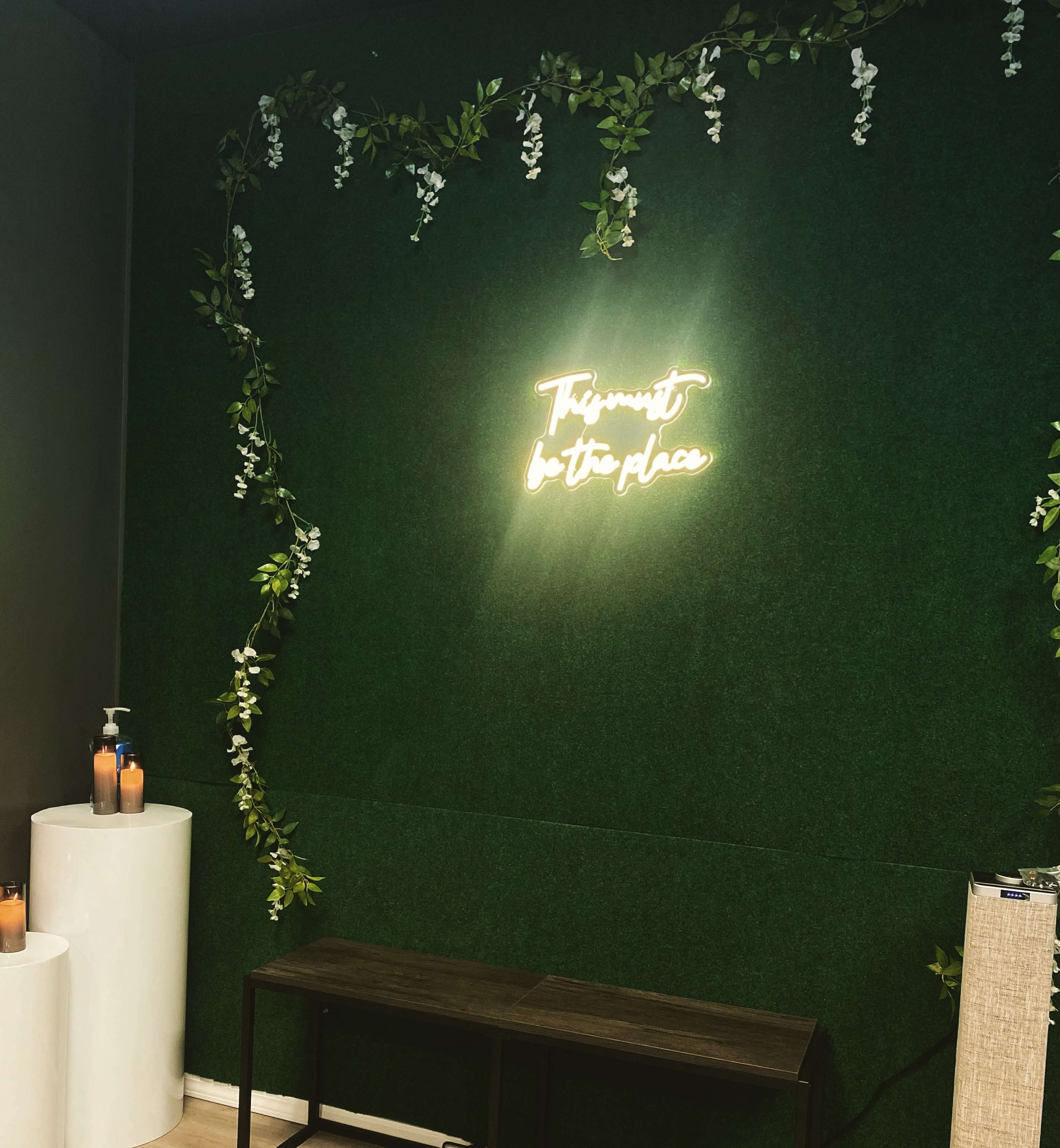 A neon sign that reads "This must be the place" is illuminated against a green wall adorned with artificial vines and floral decorations.