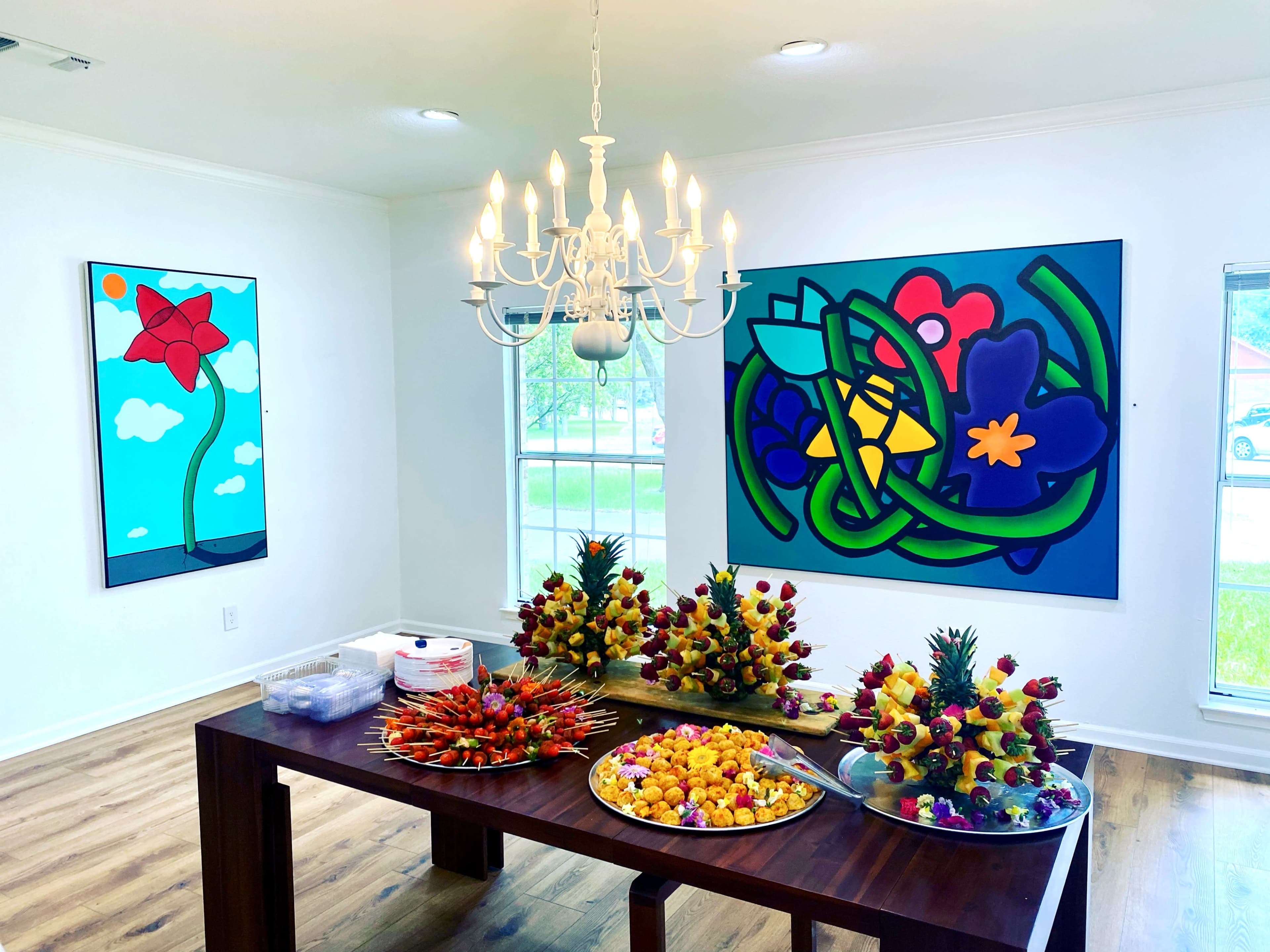 The image shows a brightly lit room featuring two colorful abstract paintings on the walls, a chandelier overhead, and a table with several fruit arrangements and platters of assorted fruit.