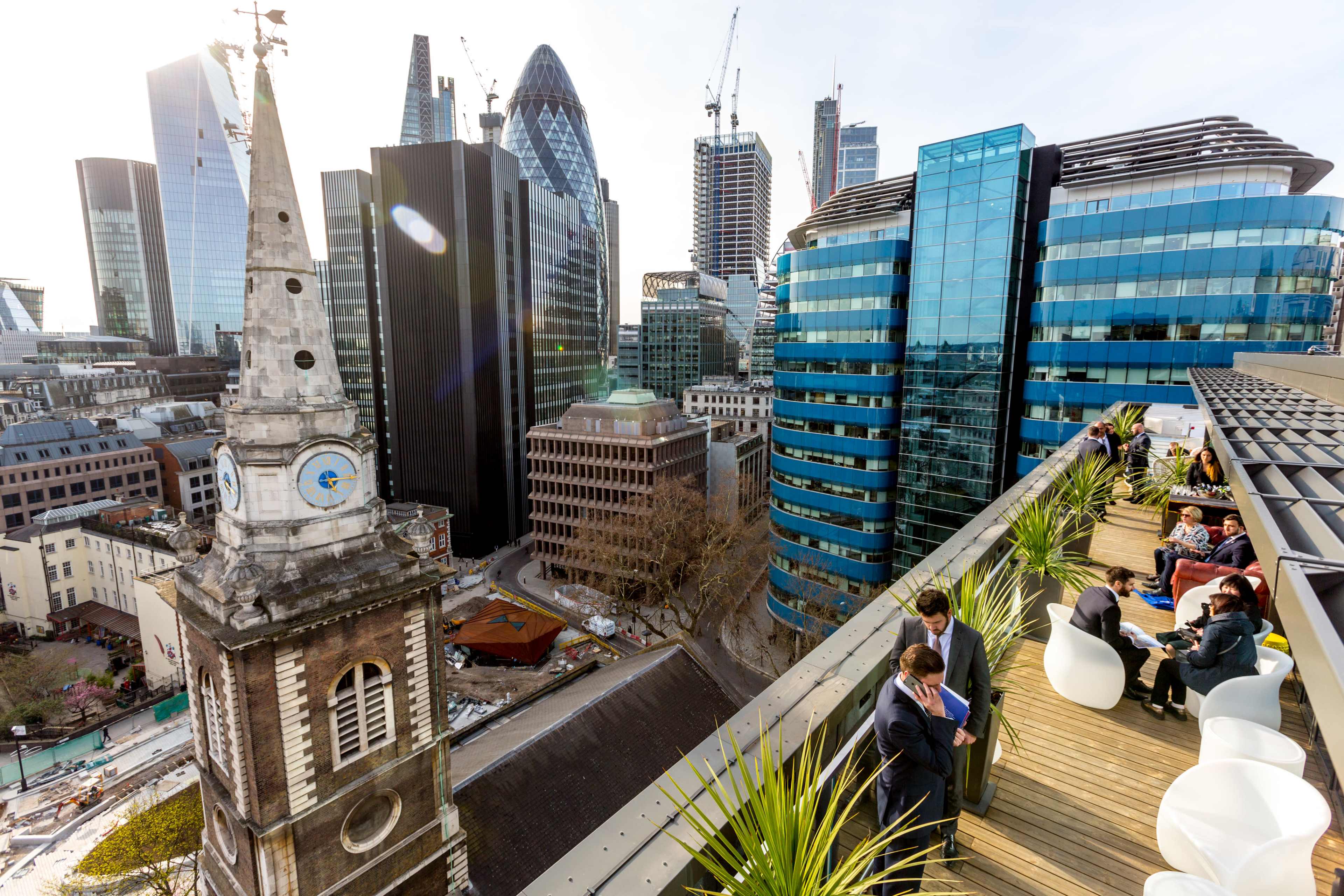 The image shows a rooftop terrace in a modern cityscape, featuring contemporary skyscrapers and a historic clock tower below.