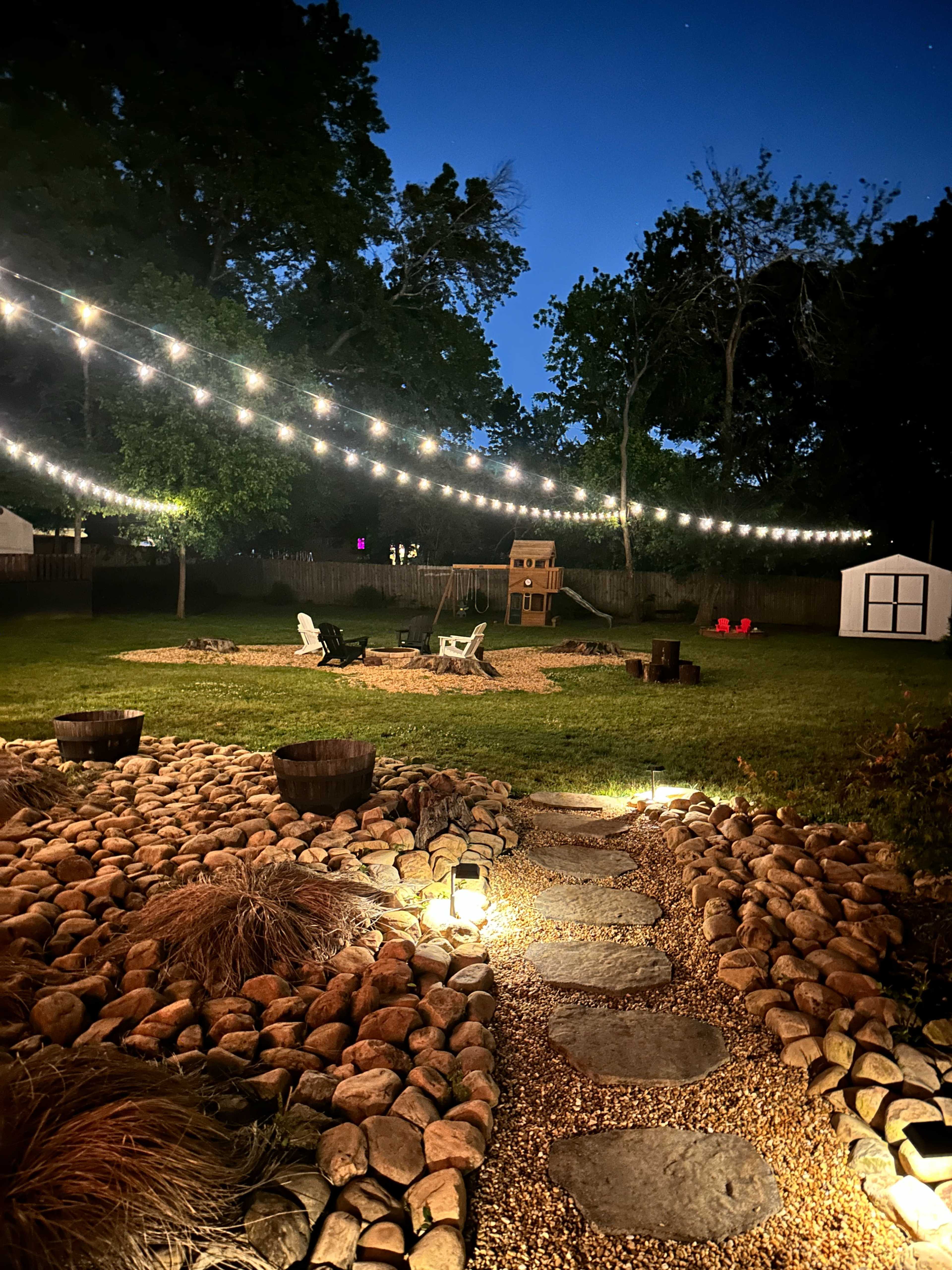 The image shows a backyard in the evening, illuminated by string lights, with a stone path leading to a gravel area featuring chairs and a playhouse.