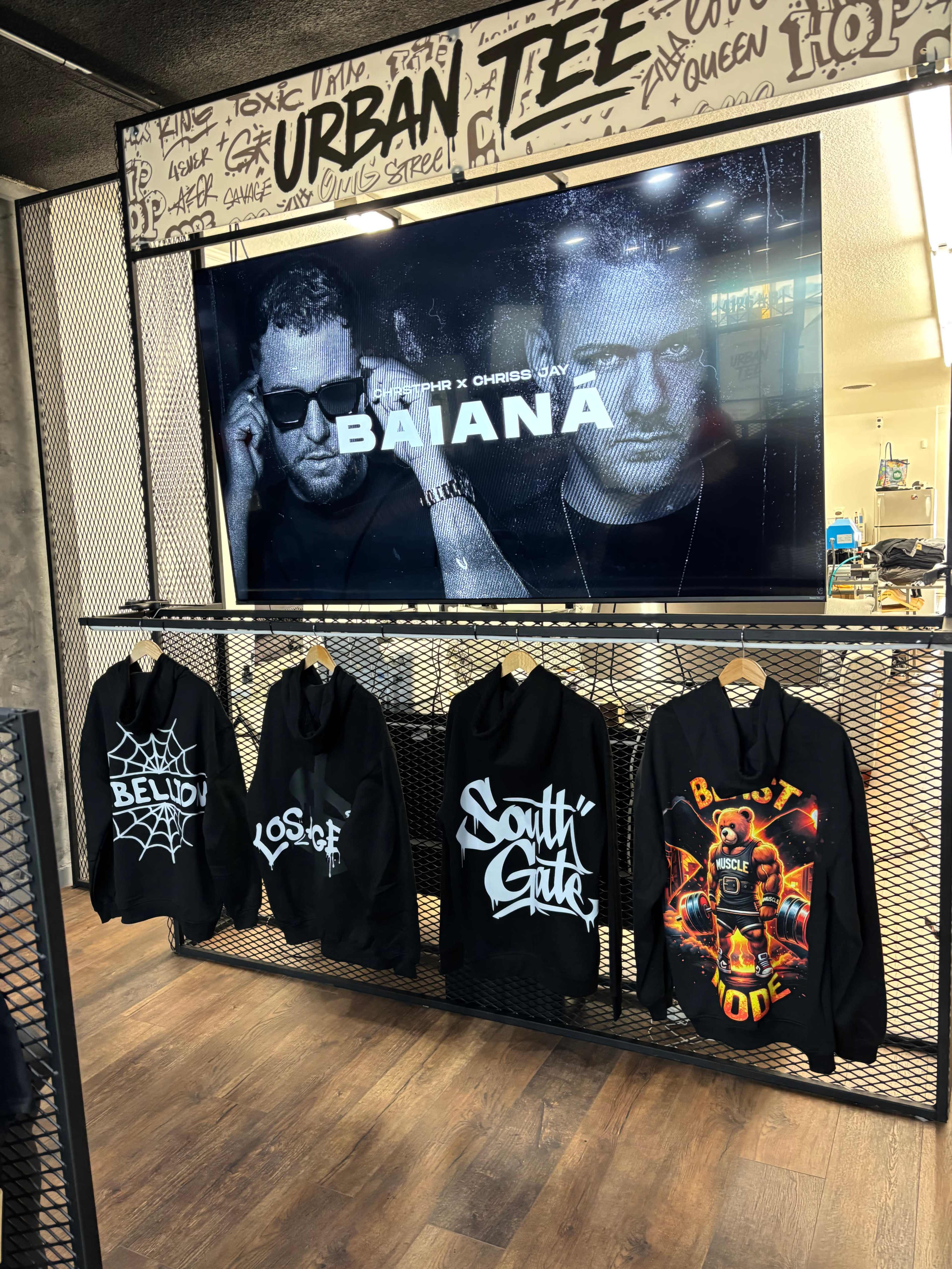 A display rack features various black hoodies with distinct designs, positioned under a large screen advertising a collaboration titled "BAIANA."