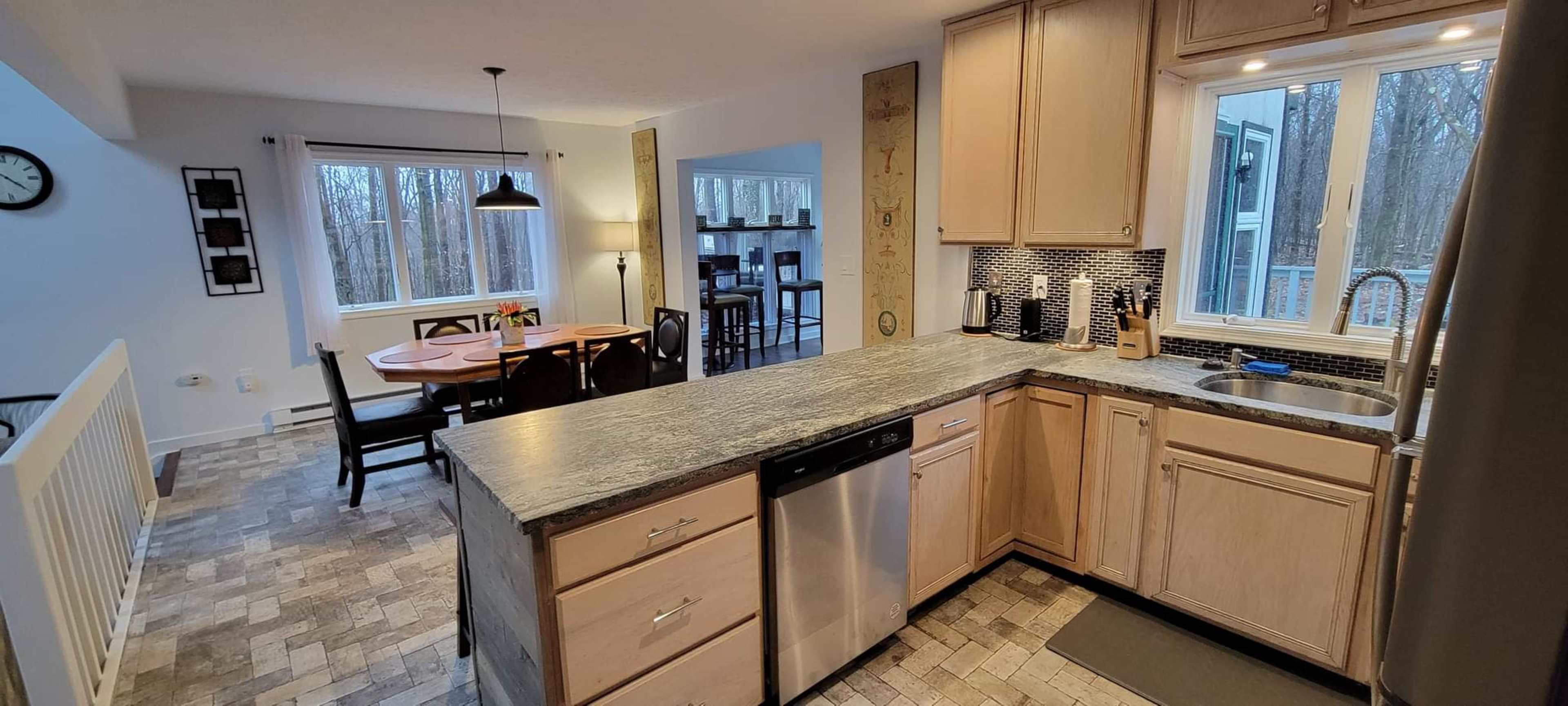The image shows a modern kitchen with light wood cabinetry, a stainless steel dishwasher, and a view of an adjoining dining area with a round table and chairs.