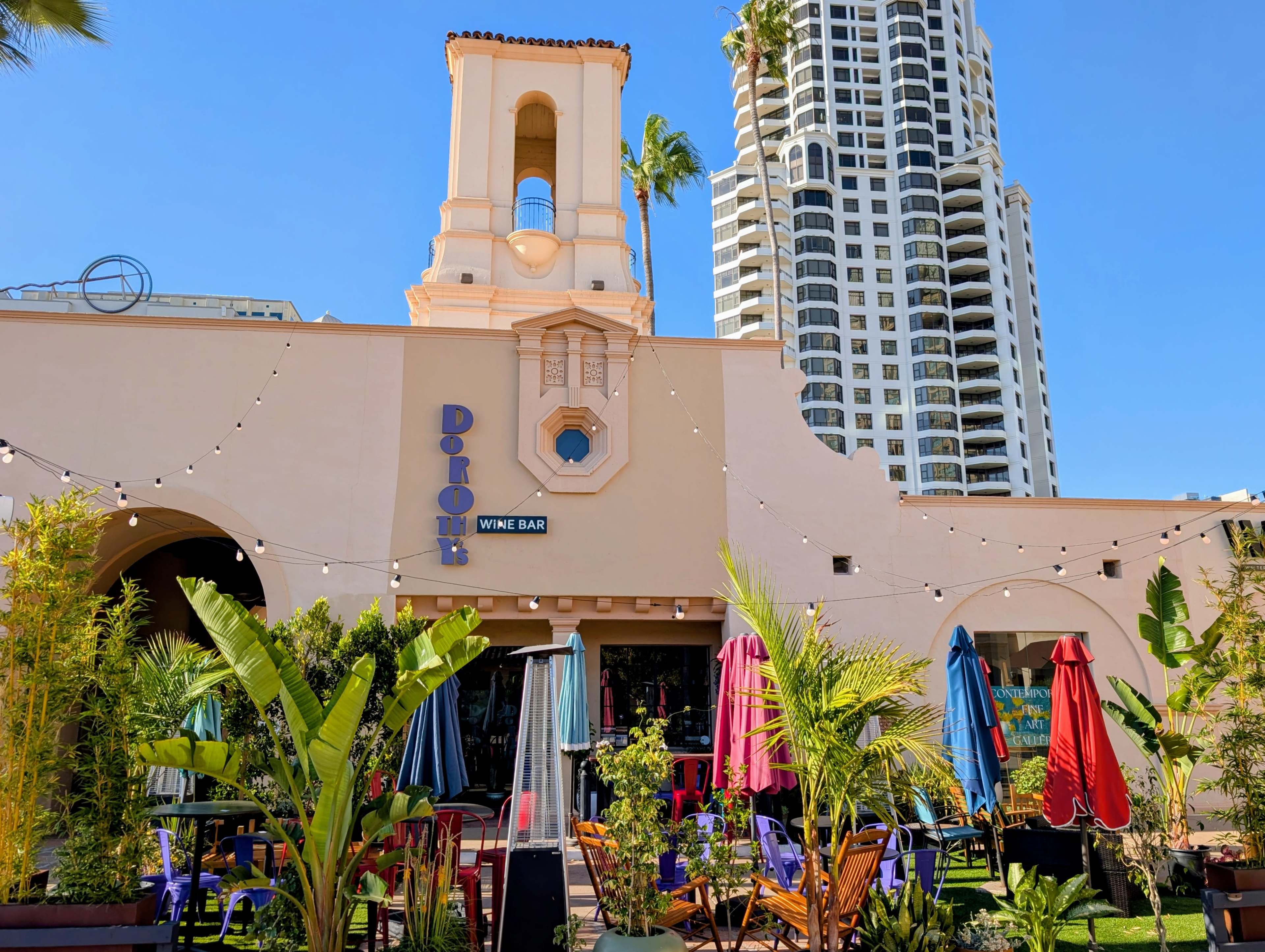 The image shows a wine bar with outdoor seating, surrounded by palm trees and colorful umbrellas, against a backdrop of tall buildings.