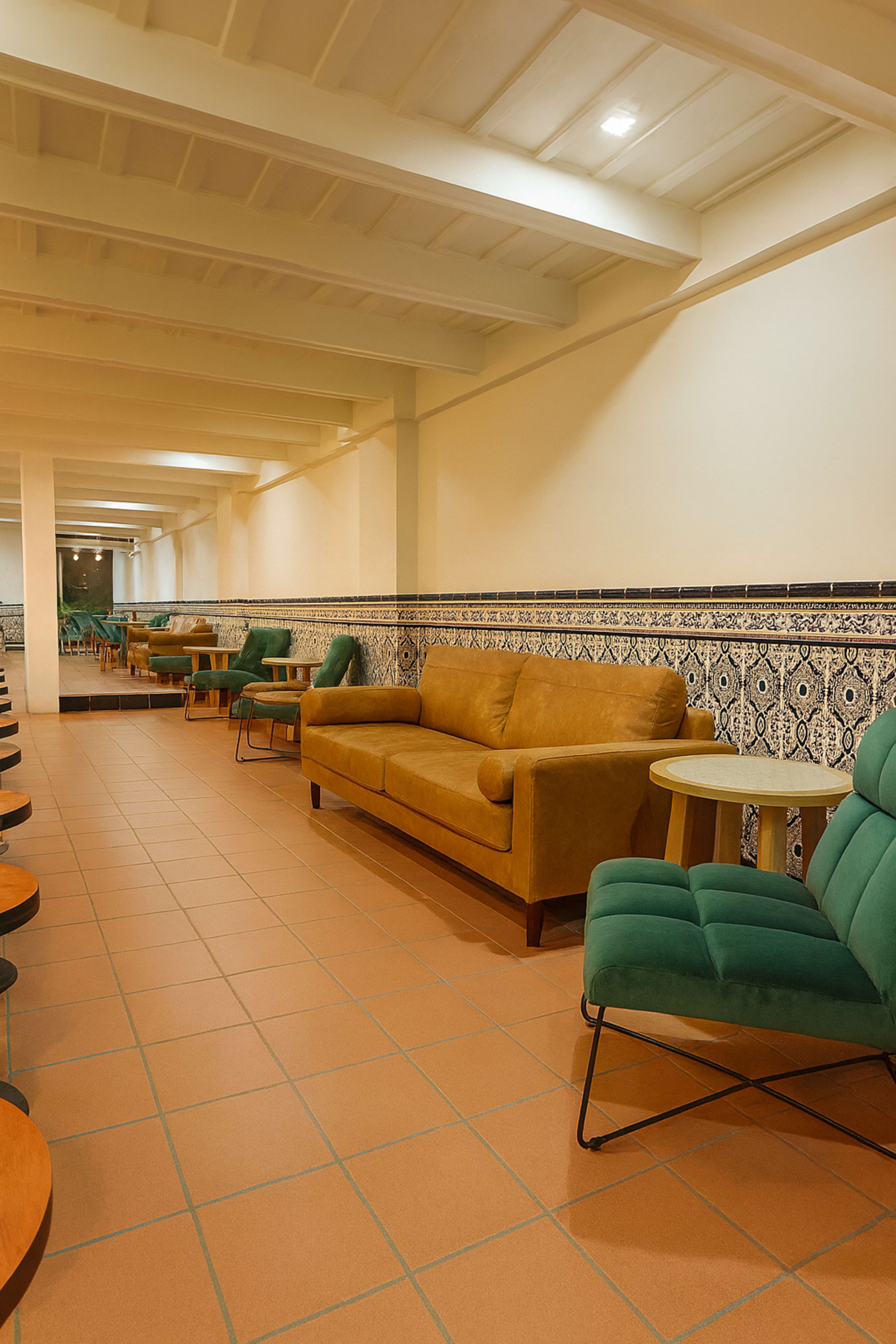 The image shows a spacious room with wooden seating areas and a soft brown couch along one wall, adorned with patterned tiles.