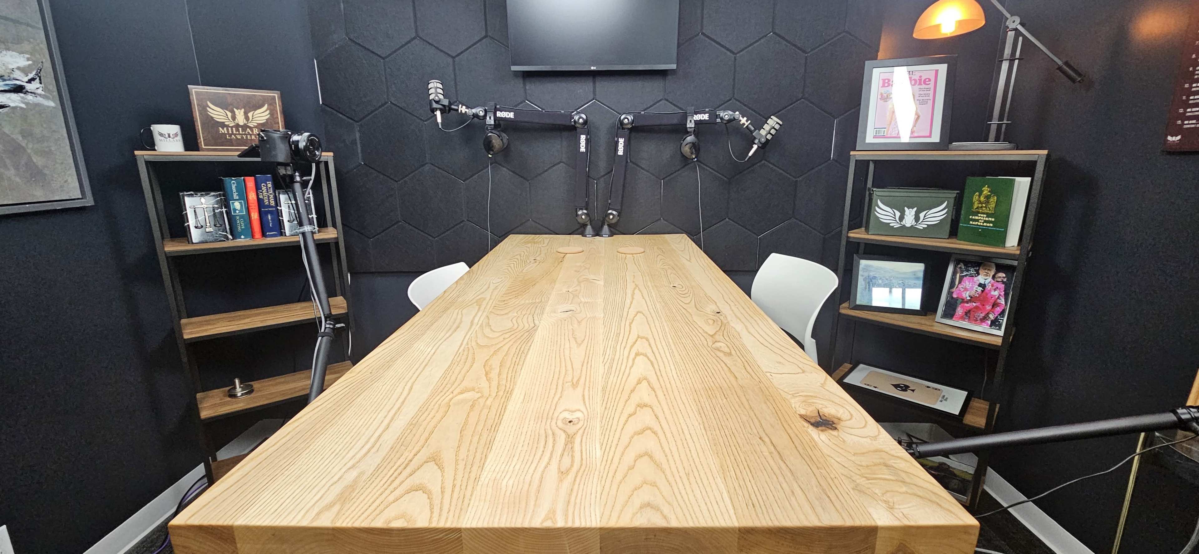 The image shows a modern recording studio with a large wooden table, two microphones mounted on adjustable arms, and bookshelves filled with books and framed photographs along the walls.