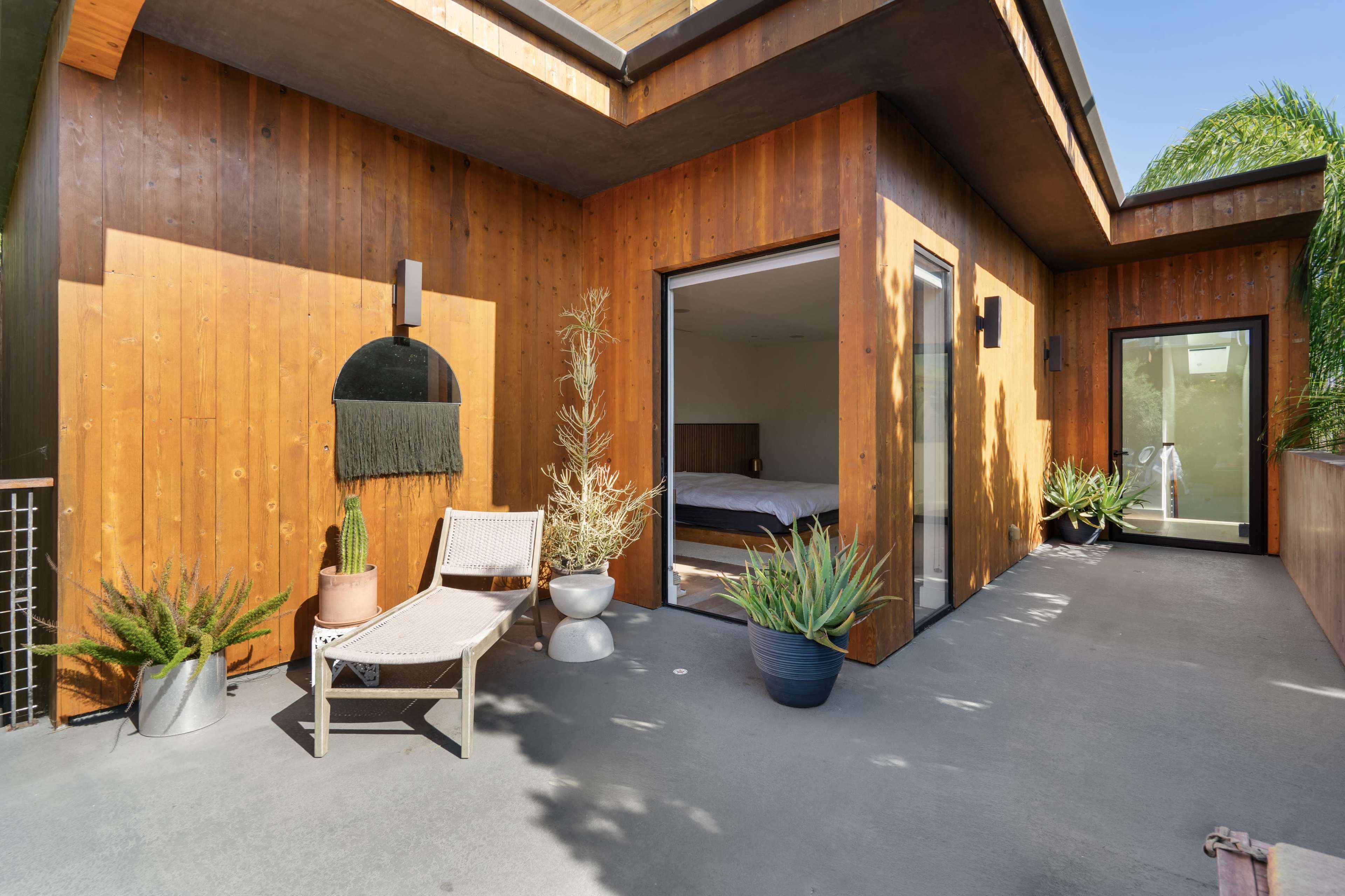 The image shows a modern patio with wooden walls, featuring seating and potted plants outside a bedroom with large glass doors.