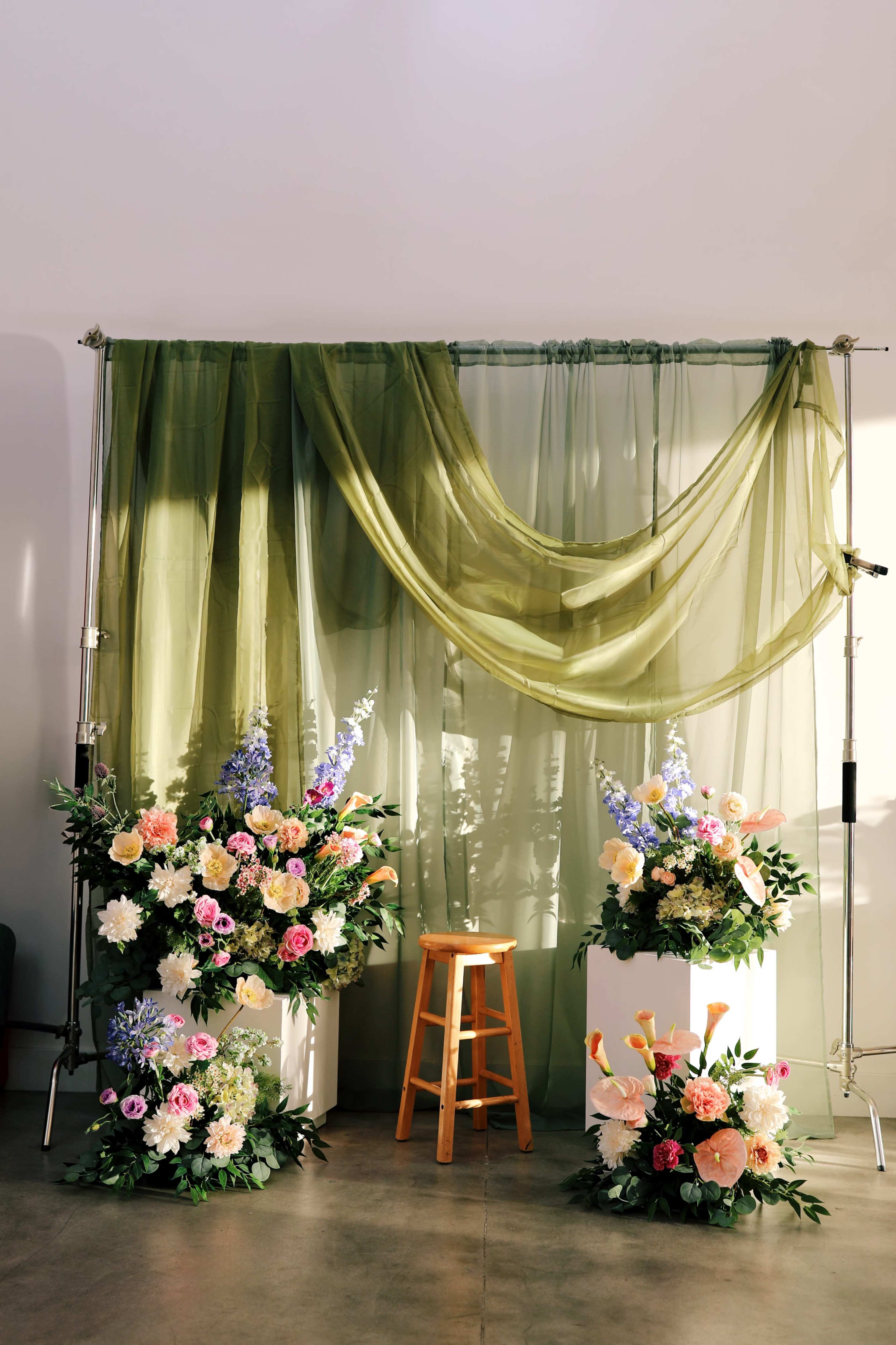 The image shows a floral arrangement with large bouquets on either side of a wooden stool, set against a backdrop of draped green fabric.