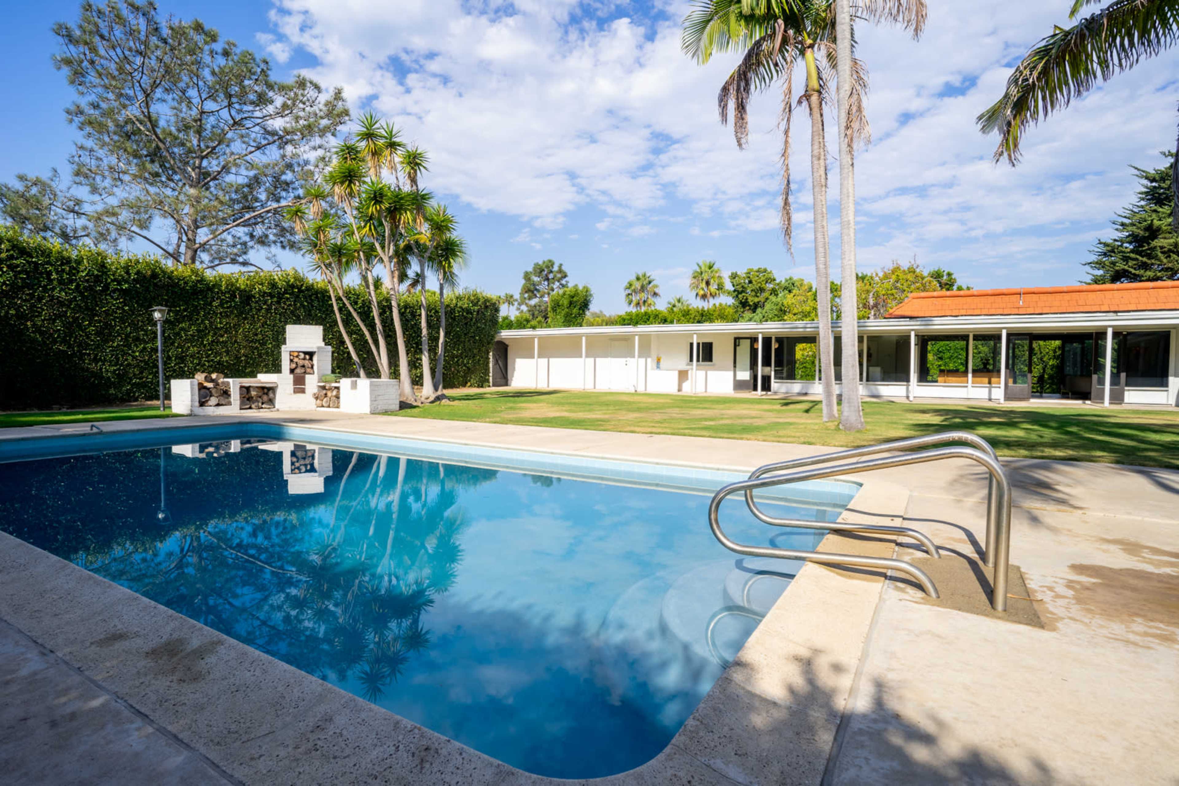 A clear swimming pool surrounded by a manicured lawn and palm trees, with a house and outdoor barbecue area visible in the background.