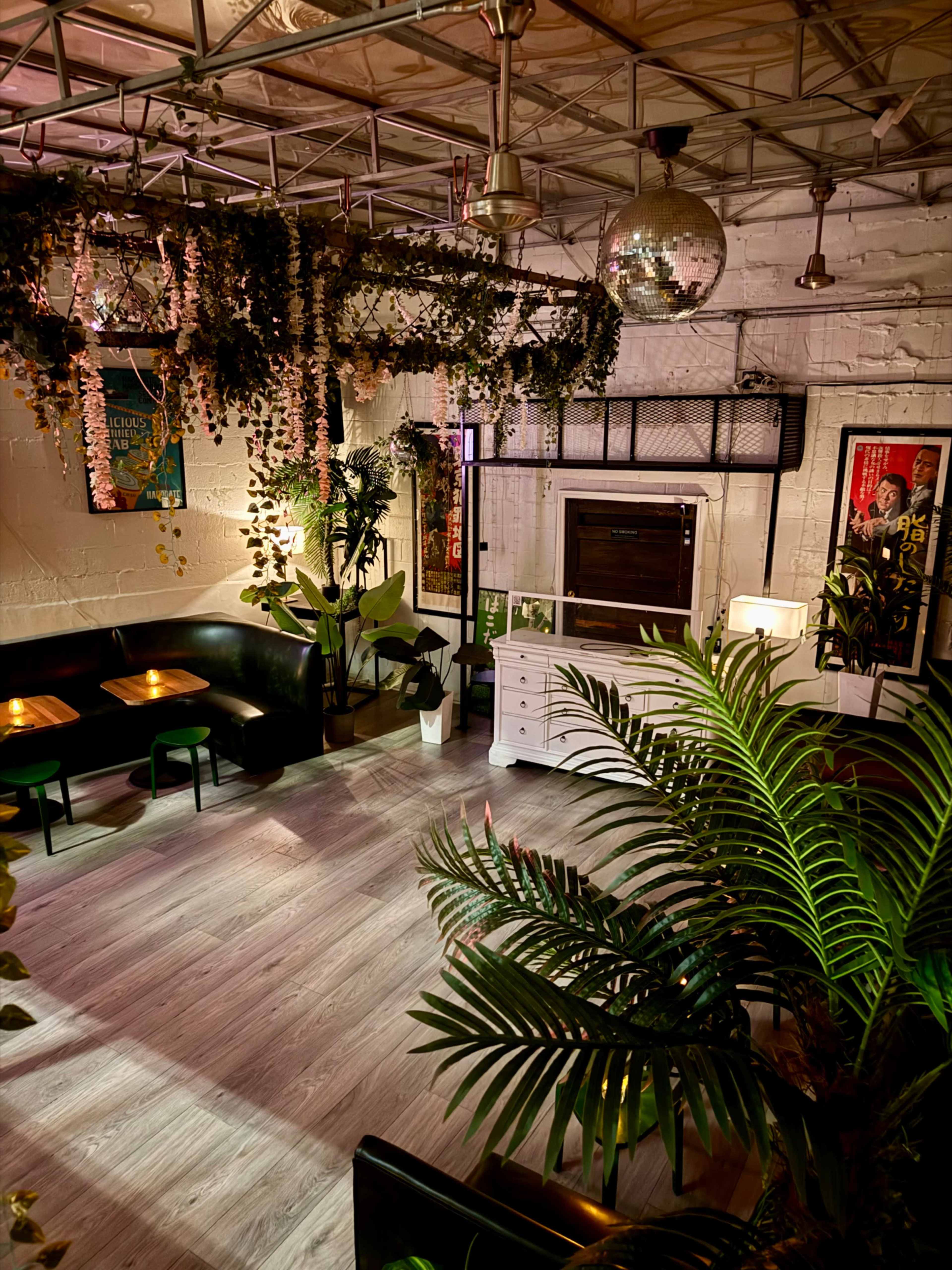 The image shows an indoor space with a modern aesthetic, featuring greenery, a disco ball, and seating areas illuminated by soft light.