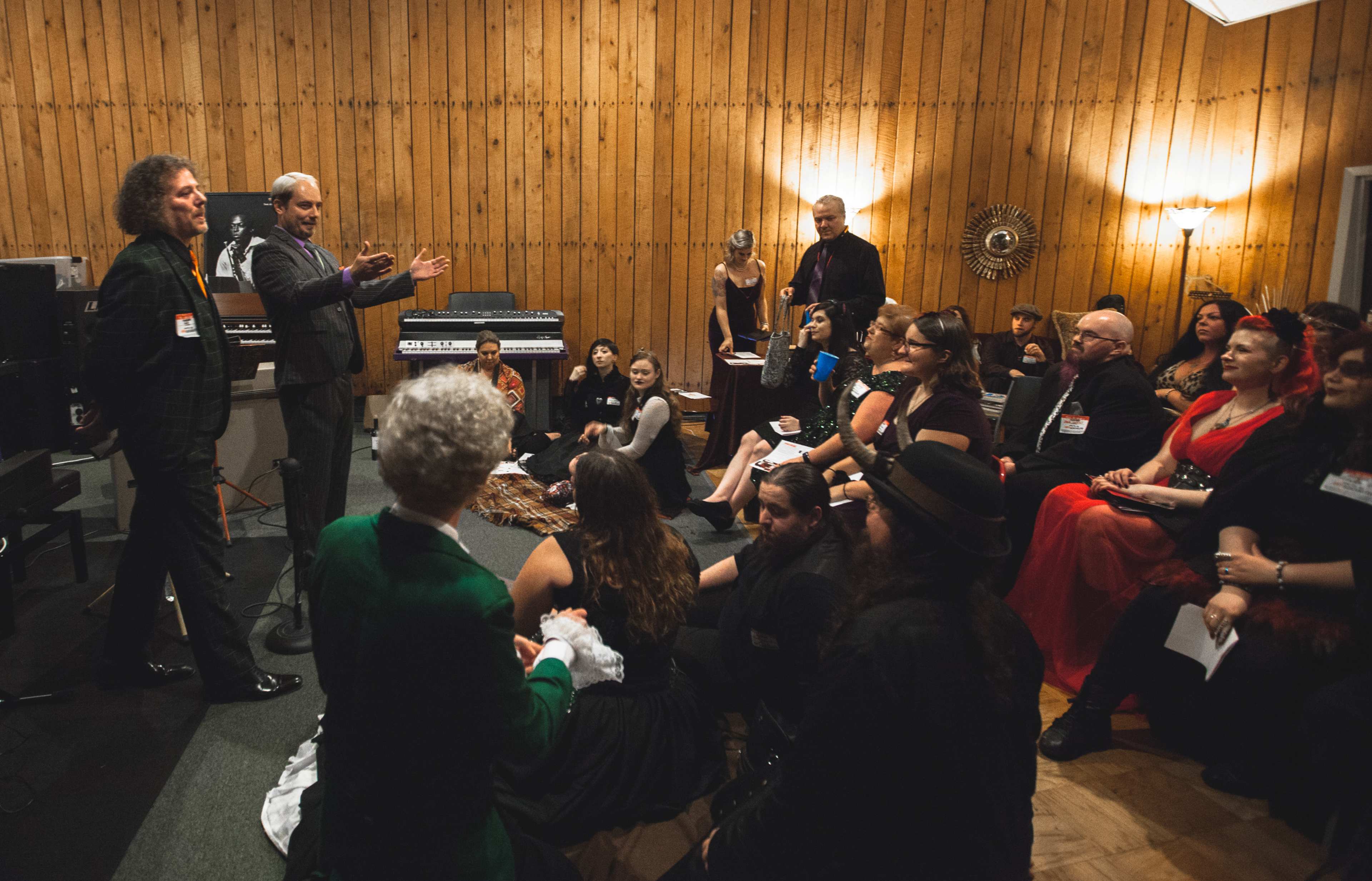 A group of people dressed in formal attire listens as two men speak in a room with wooden paneling and musical instruments.
