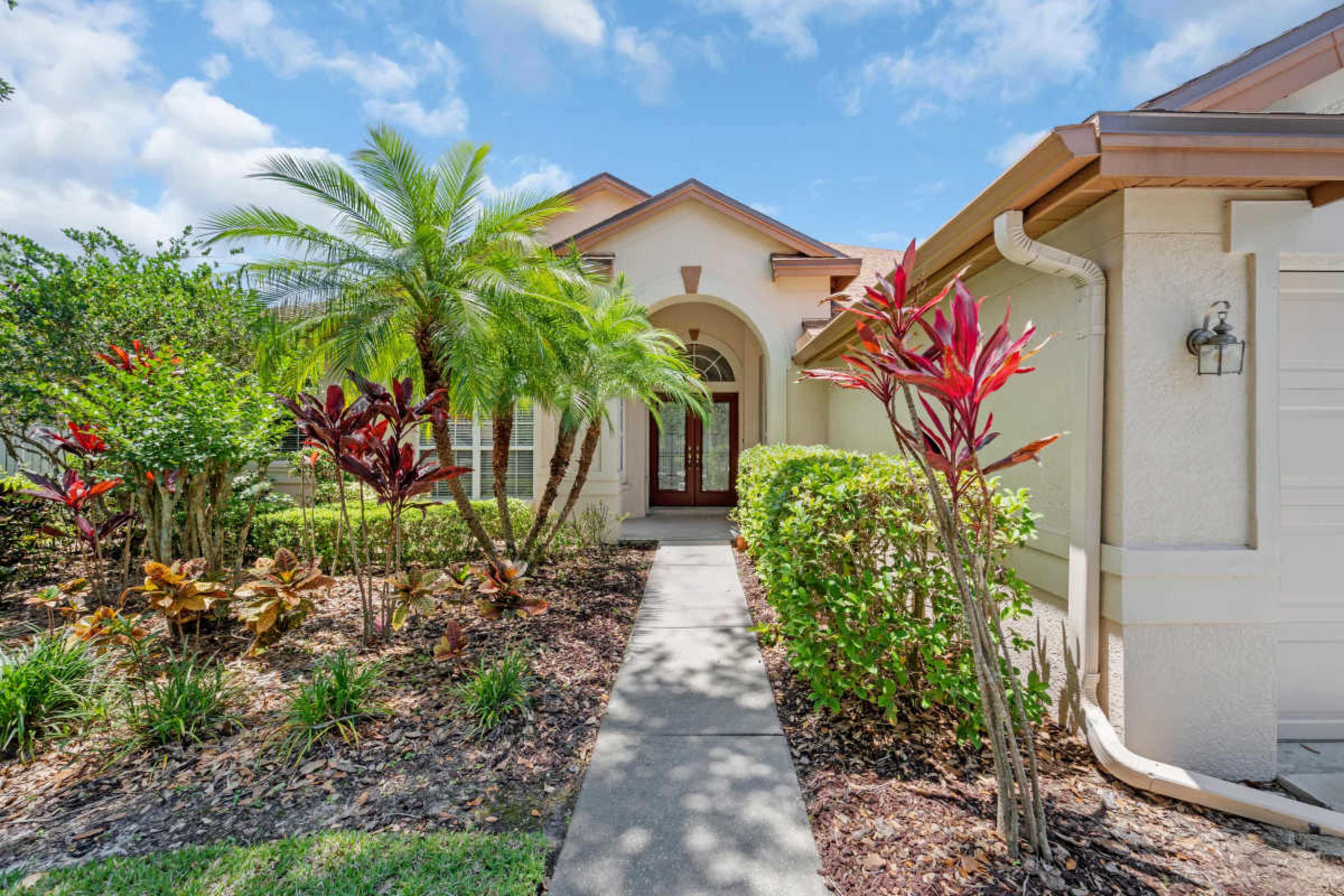 The image shows the front entrance of a house with a landscaped pathway lined by tropical plants and shrubs.