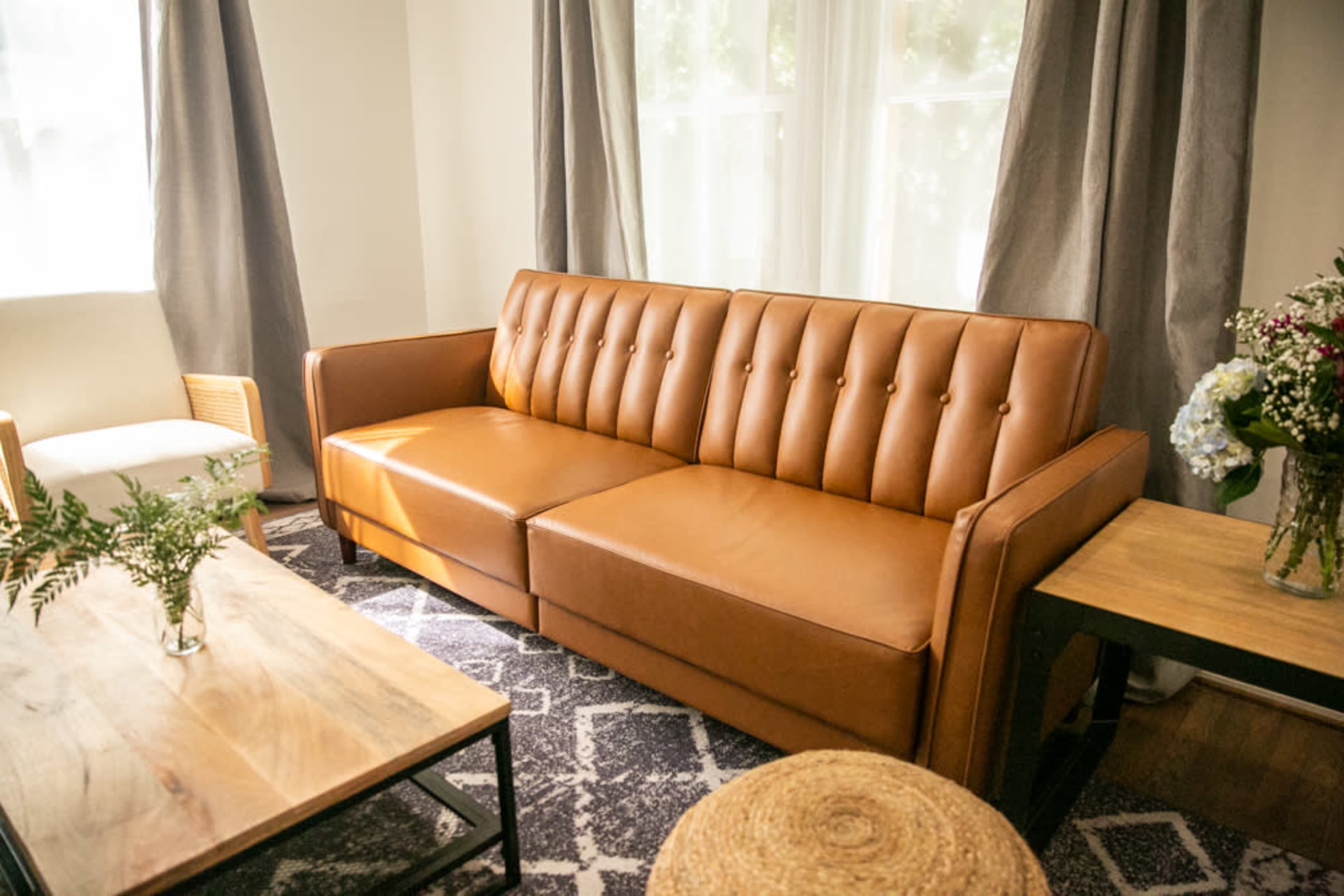 A modern living room featuring a brown leather tufted sofa, two wooden tables, and a vase of flowers, with large windows and grey curtains.