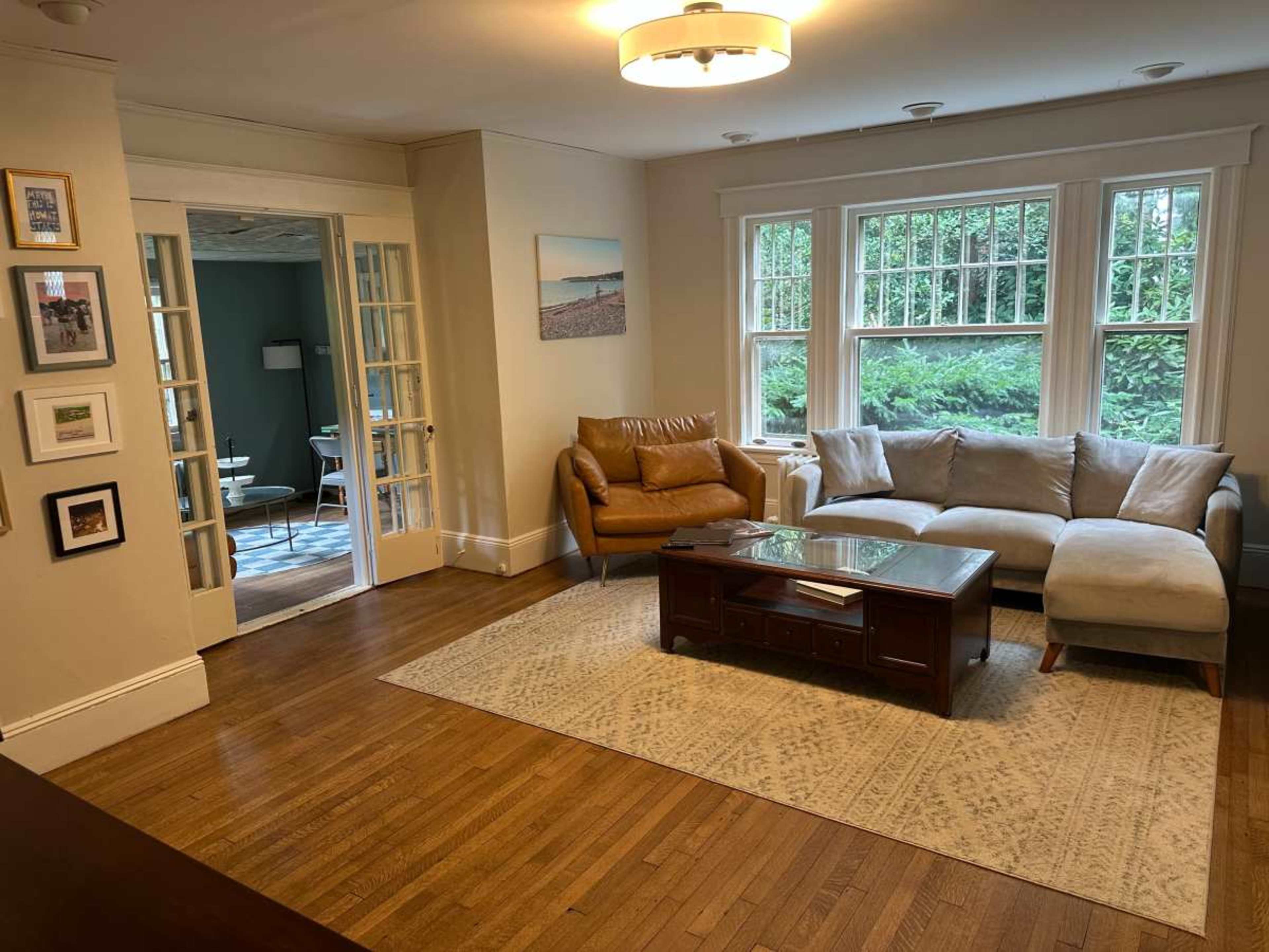 The image shows a living room with a brown leather chair, a light-colored sectional sofa, a wooden coffee table, and large windows overlooking greenery.