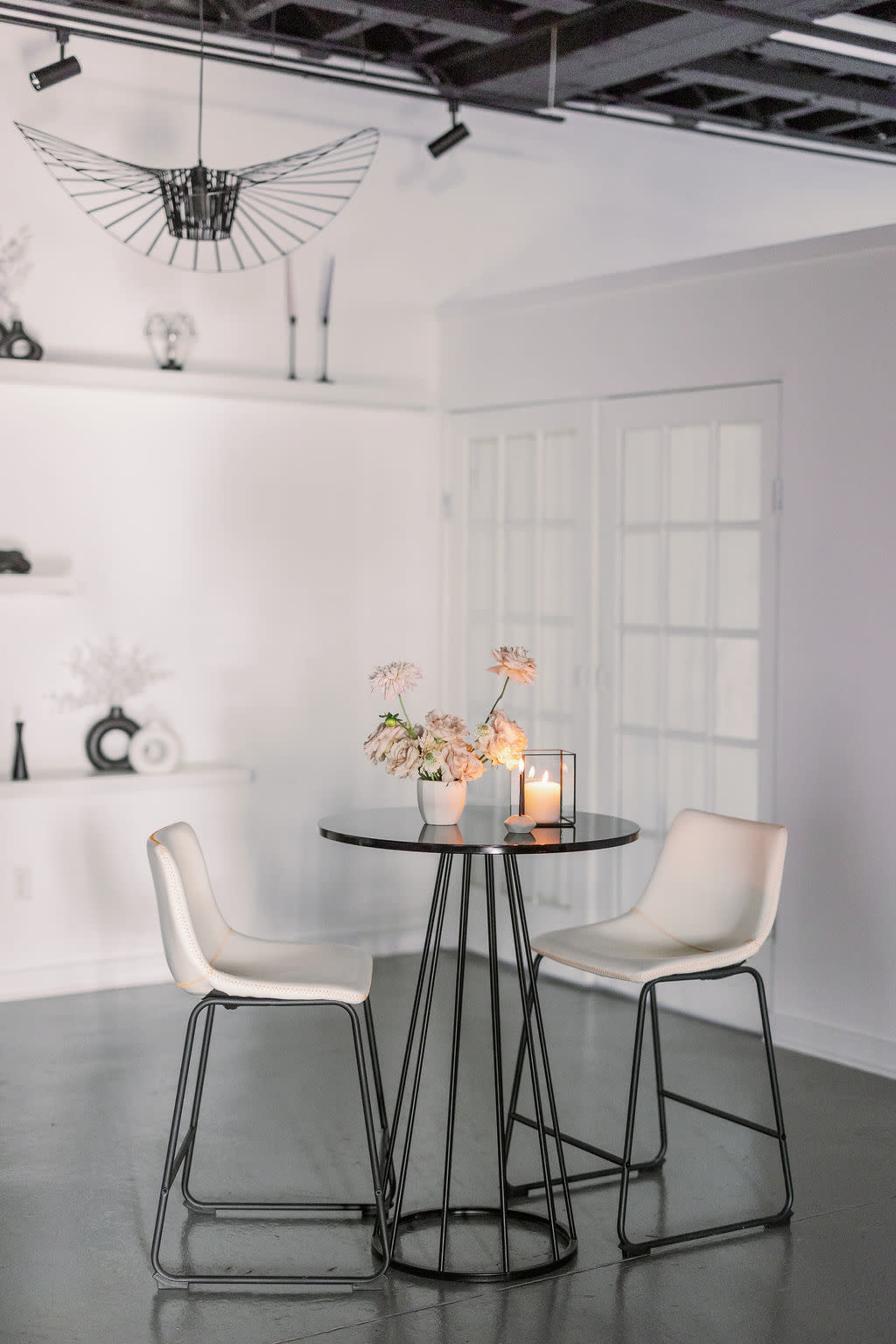 A small, modern dining table with two white chairs is placed in a minimalist room featuring a candle and floral arrangement on the table.