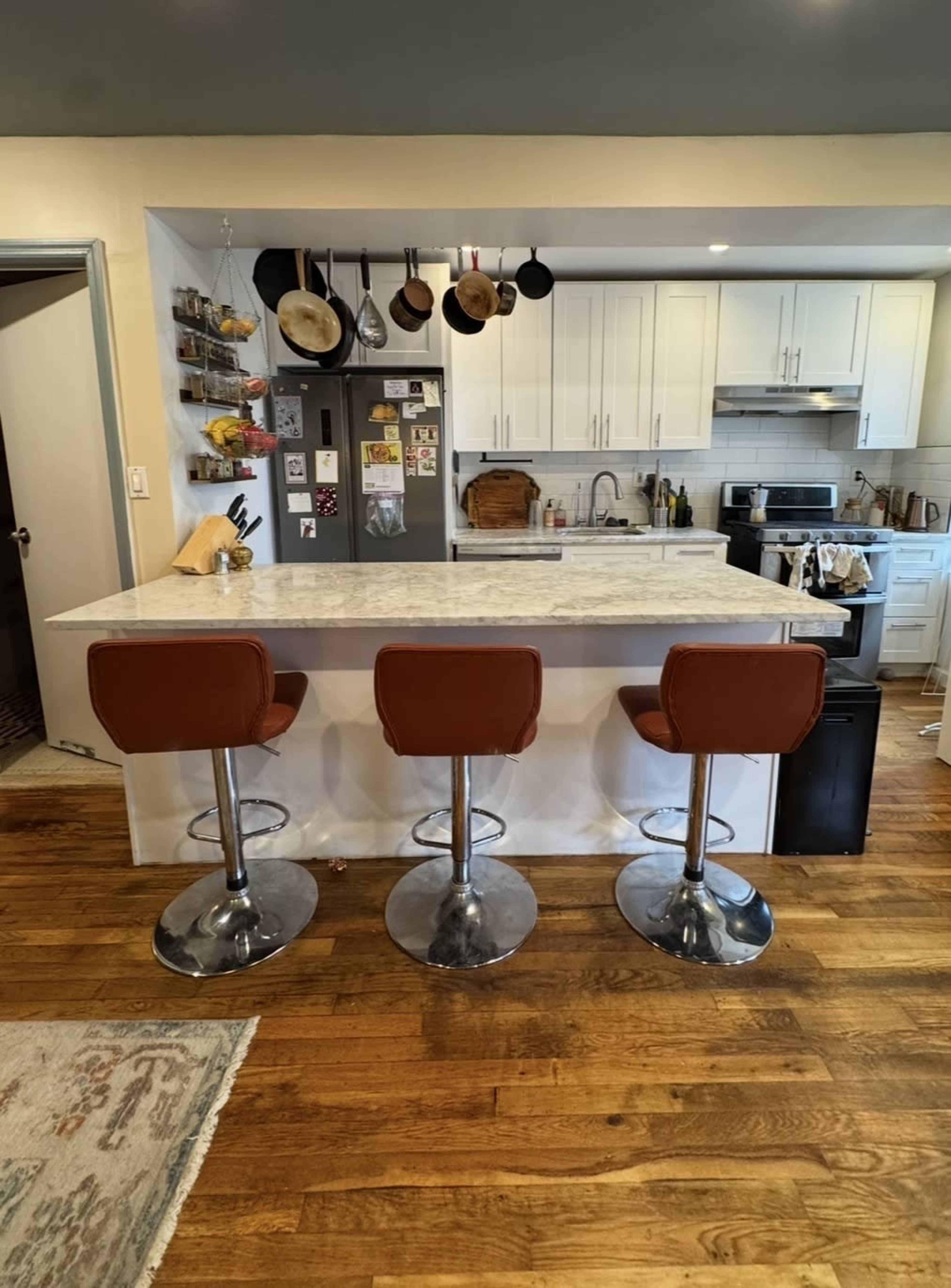 The image shows a modern kitchen with a marble countertop, three chrome bar stools, and an assortment of cooking utensils hanging on a rack above the counter.