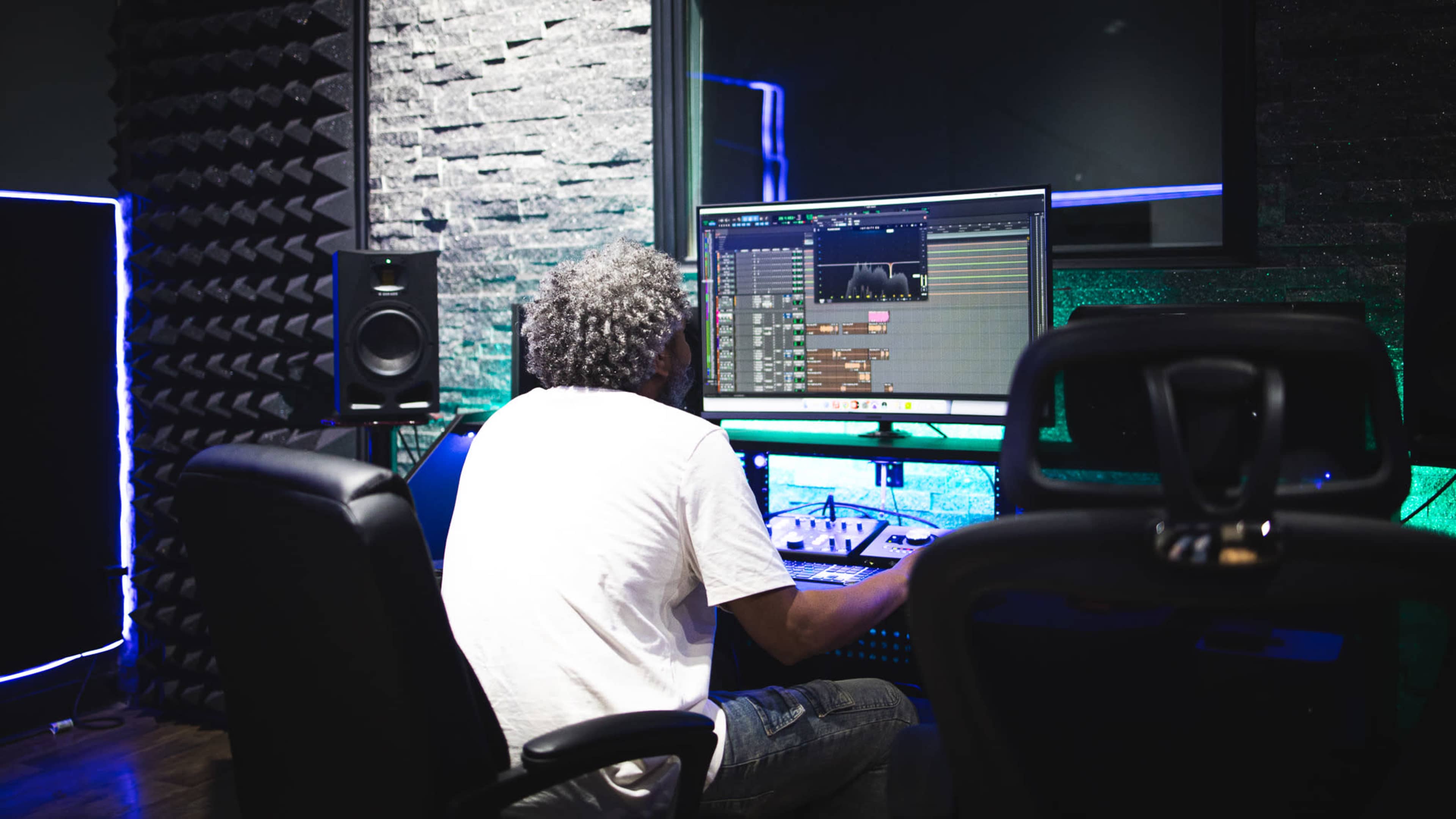 A person is working on music production in a recording studio, sitting in front of a computer with a digital audio workstation displayed on the screen.