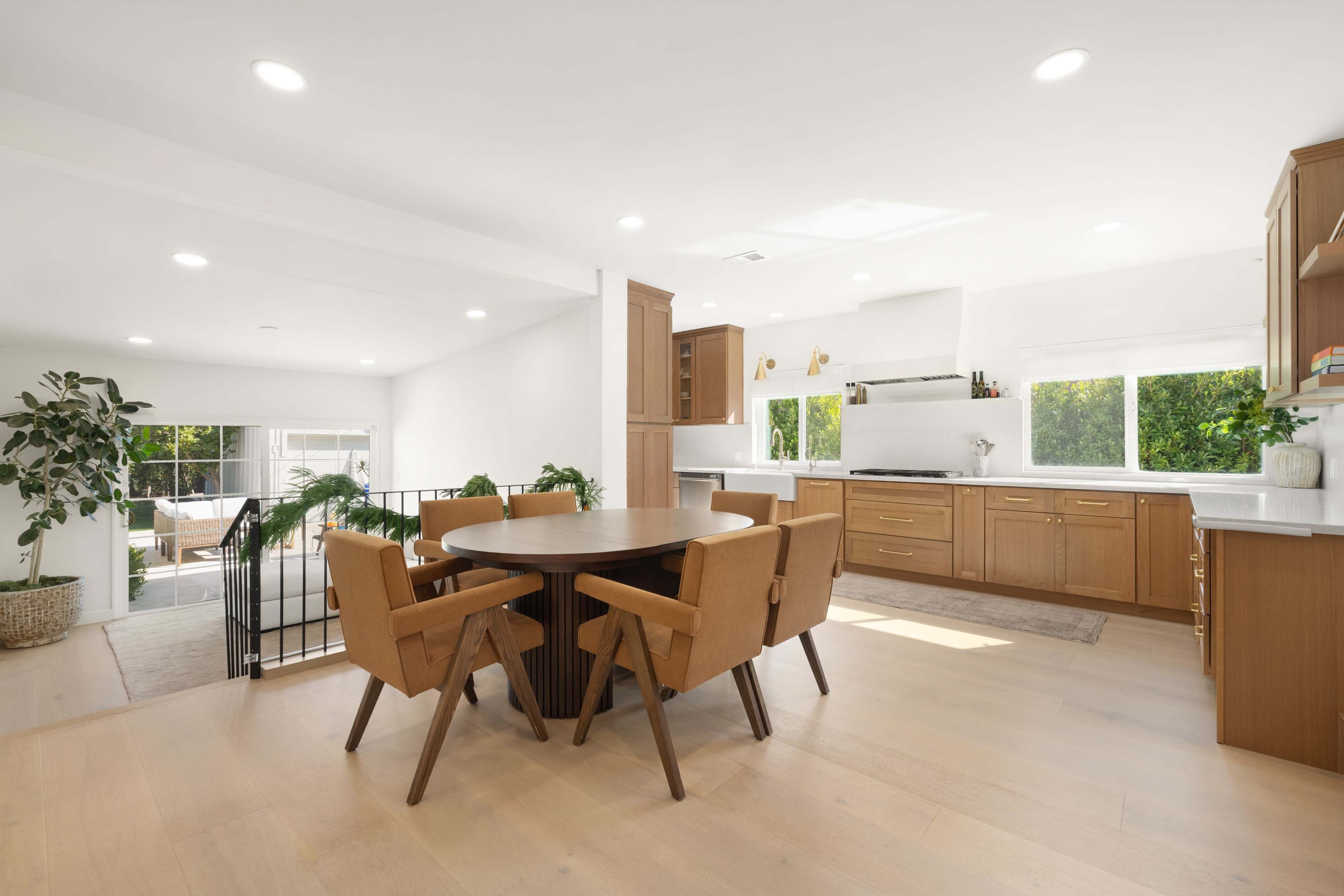 The image shows a modern kitchen-dining area with a round wooden table surrounded by four upholstered chairs, large windows allowing natural light, and sleek cabinetry.
