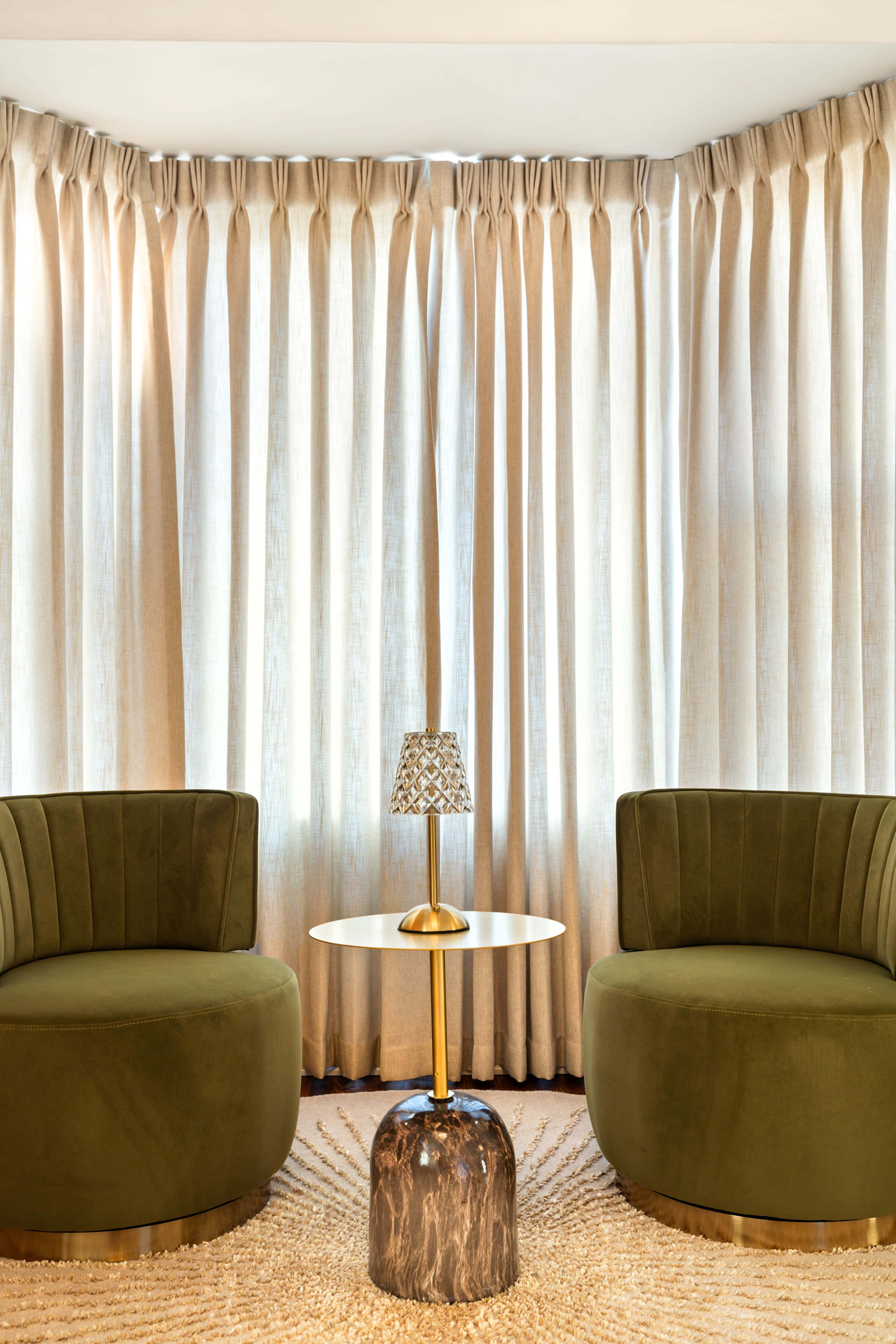 The image features two green upholstered chairs facing each other across a small round table, with light-colored curtains hanging in the background.
