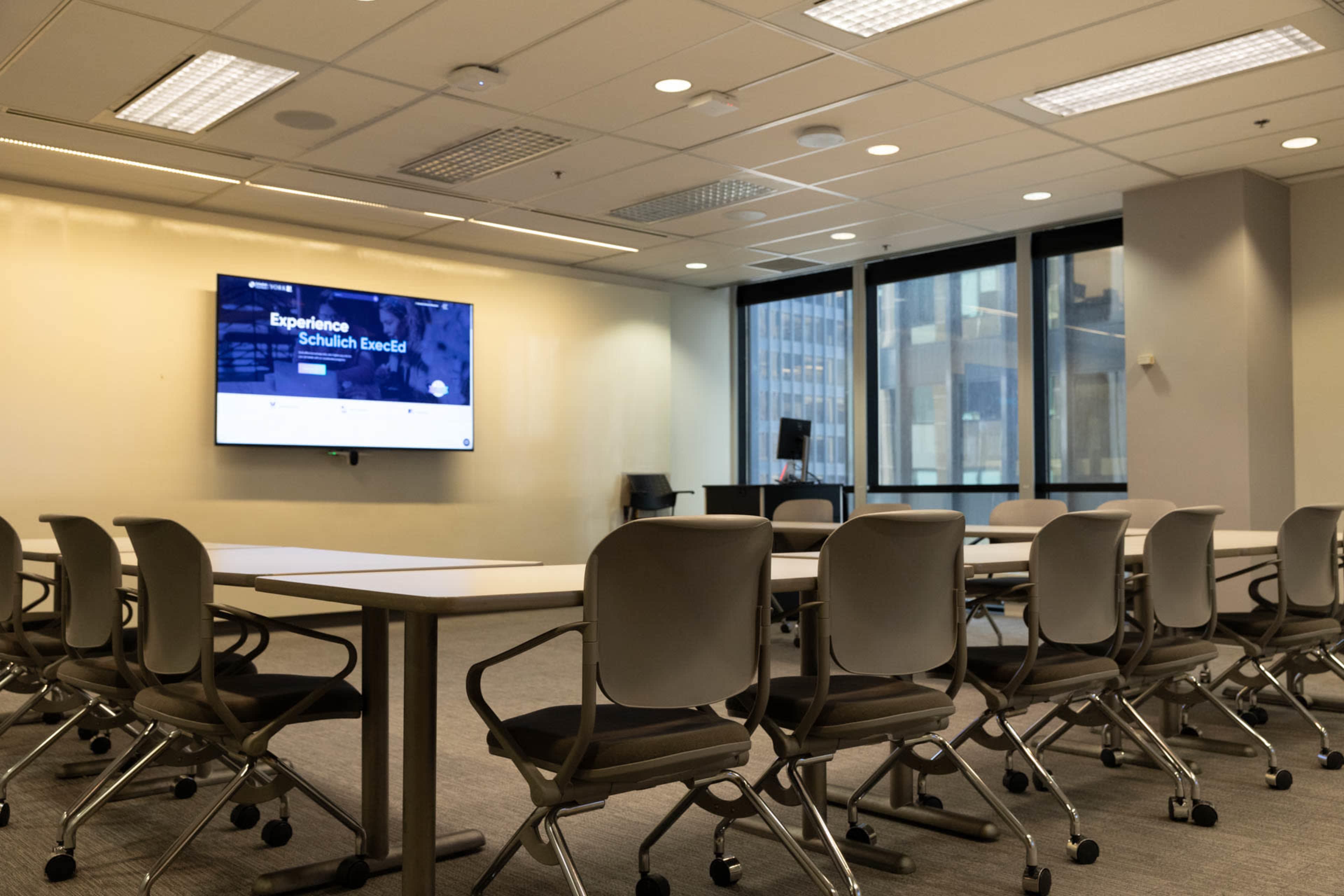 A modern conference room features a large screen on one wall, multiple tables arranged in a U-shape, and ergonomic chairs.