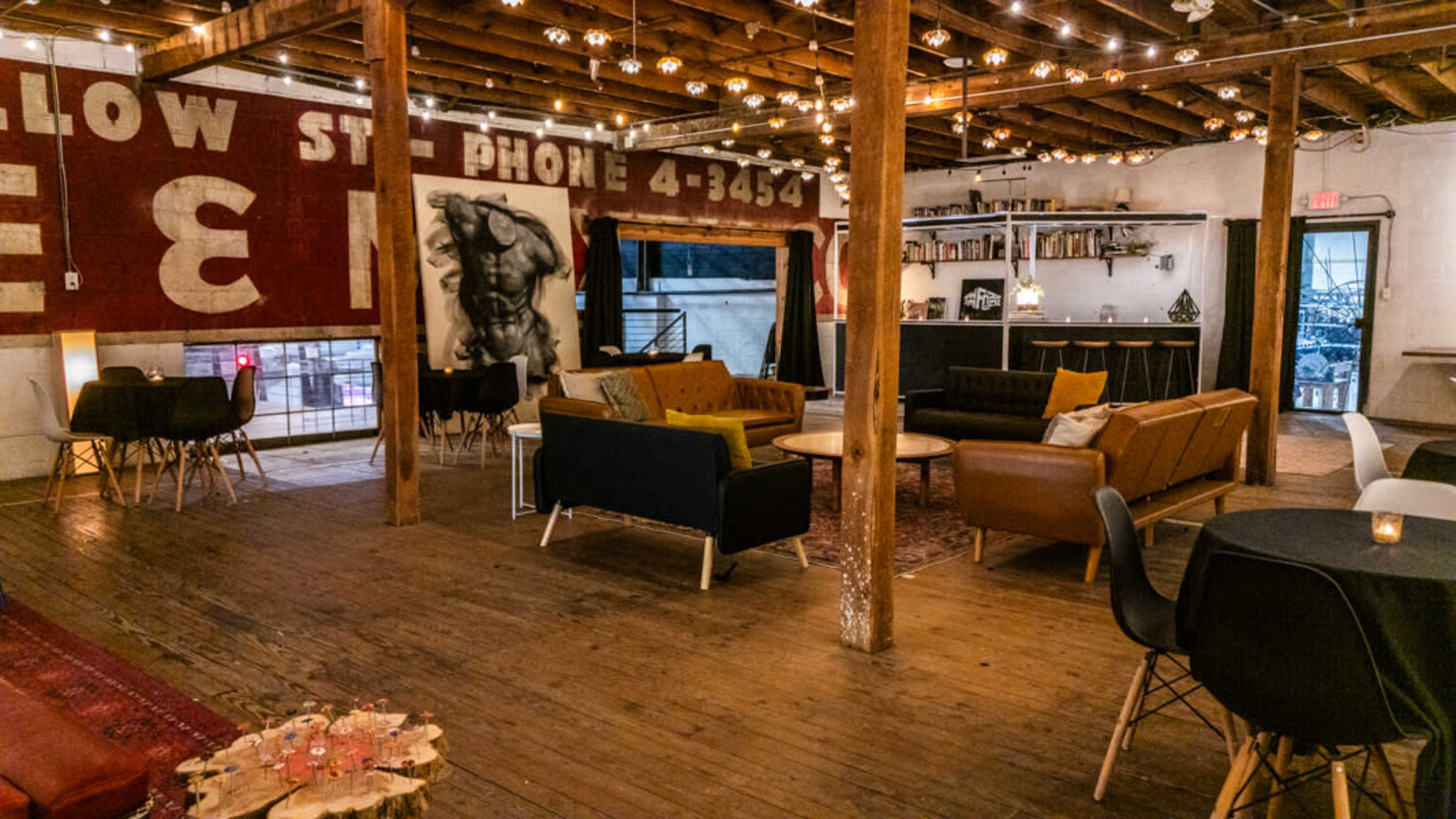 The image depicts an elegantly designed interior space with wooden beams, warm lighting, and a mix of modern furniture arranged around a central area.