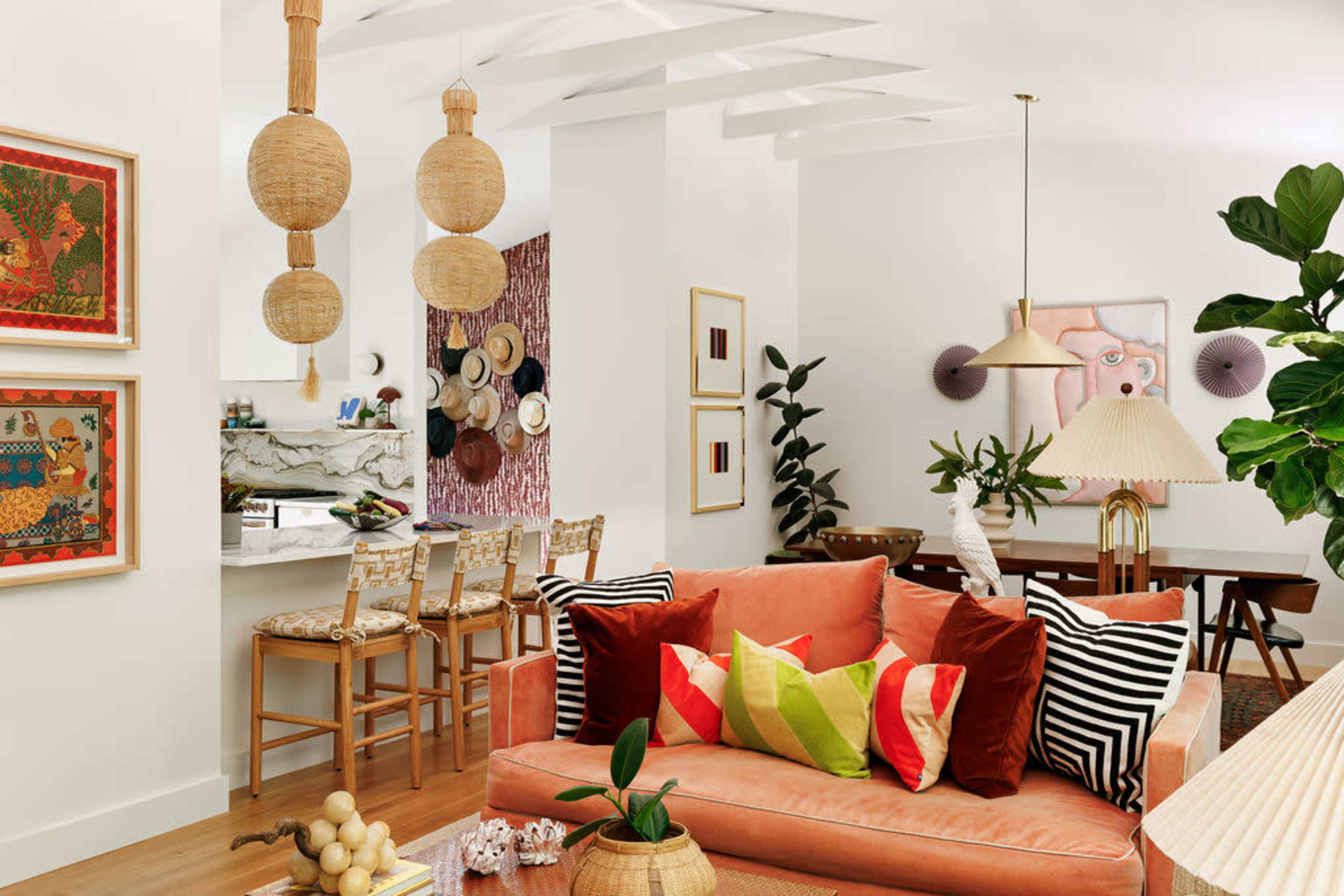 The image depicts a modern living room featuring a coral sofa adorned with colorful cushions, a dining area in the background, and decorative lighting.