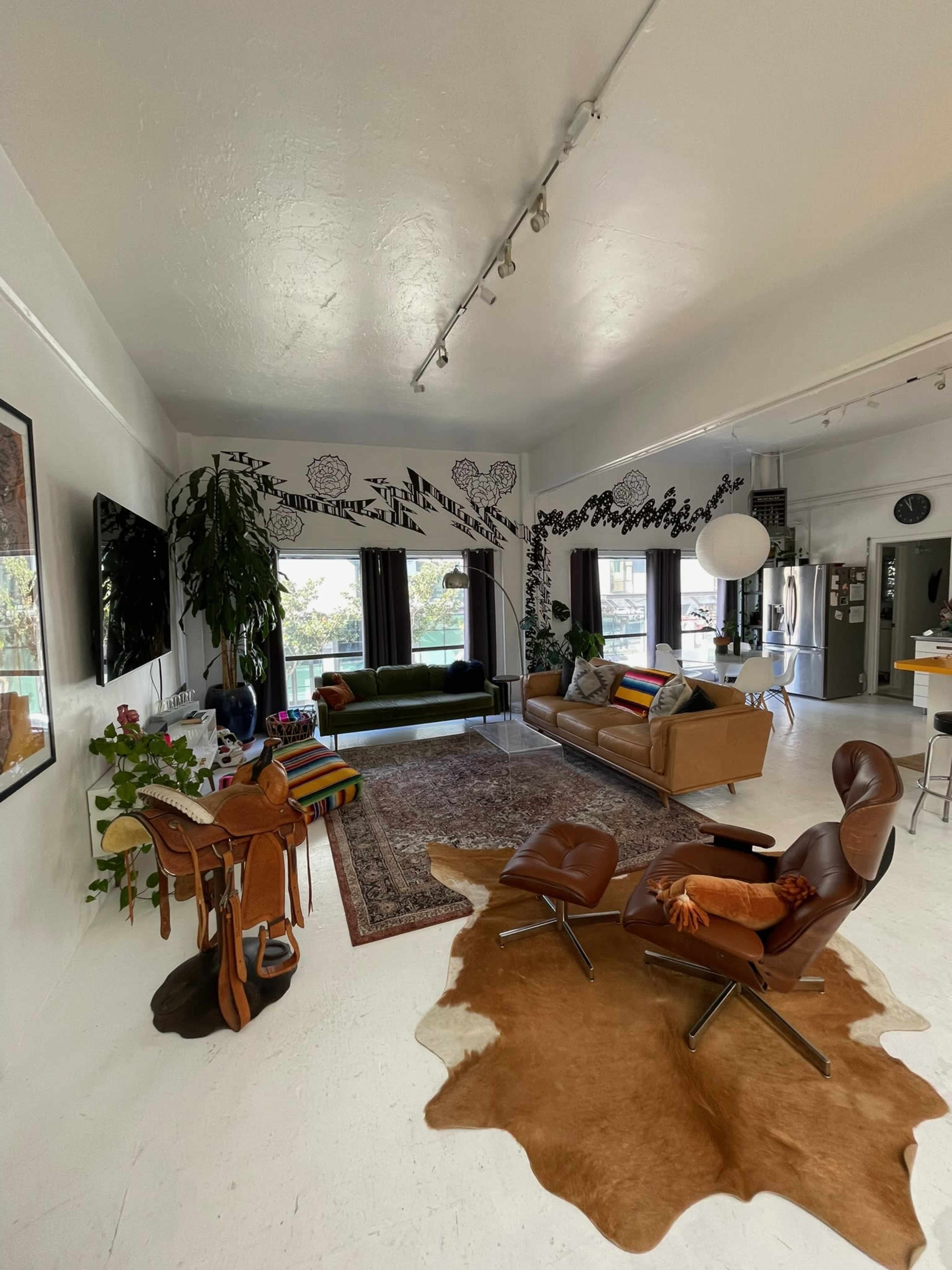 The image shows a modern living room with a large green sofa, a brown leather couch, and a central glass coffee table, surrounded by plants and artwork on the walls.