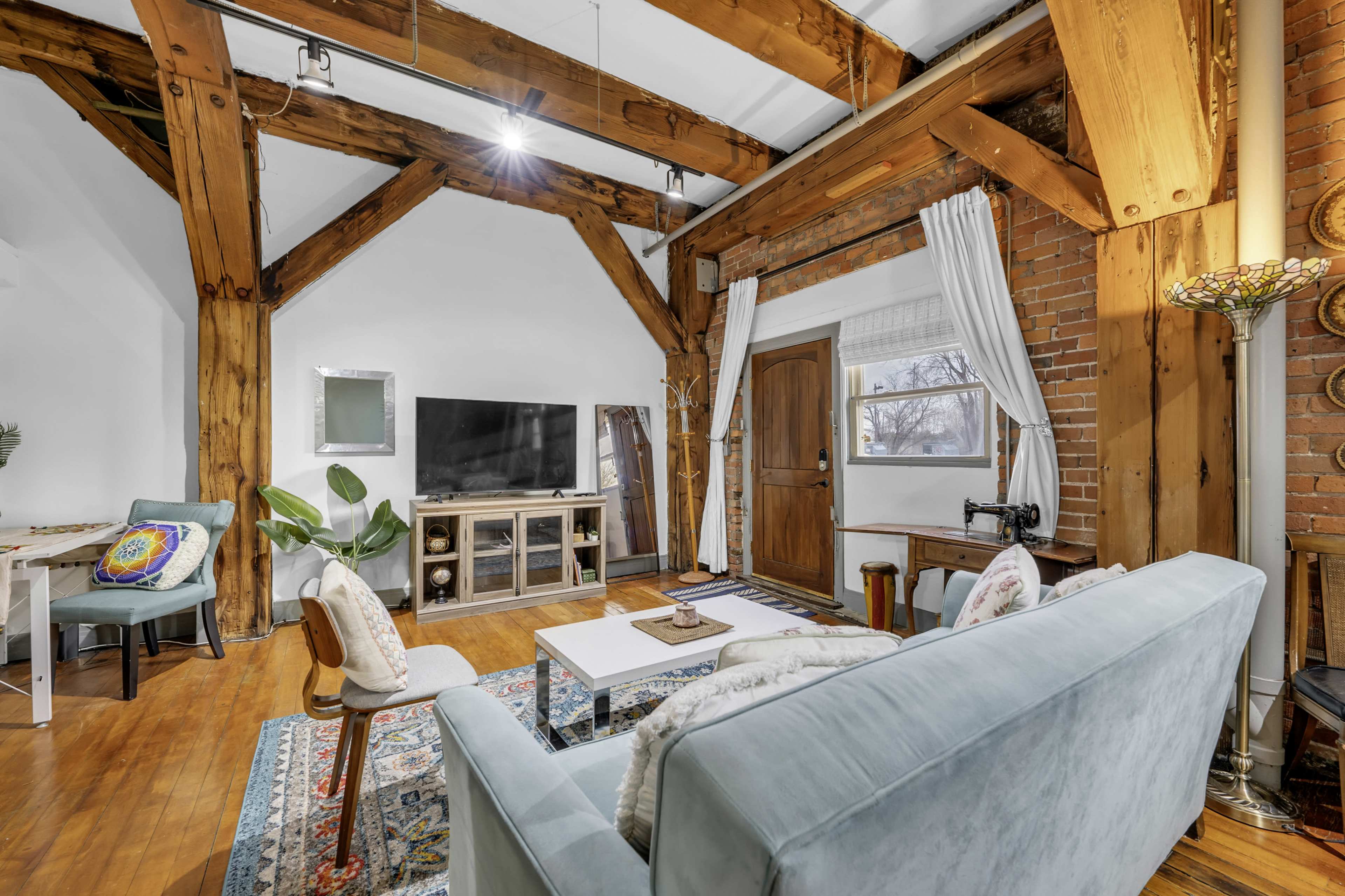 The image shows a cozy living room featuring exposed wooden beams, a television, a sewing machine, and a door leading outside.