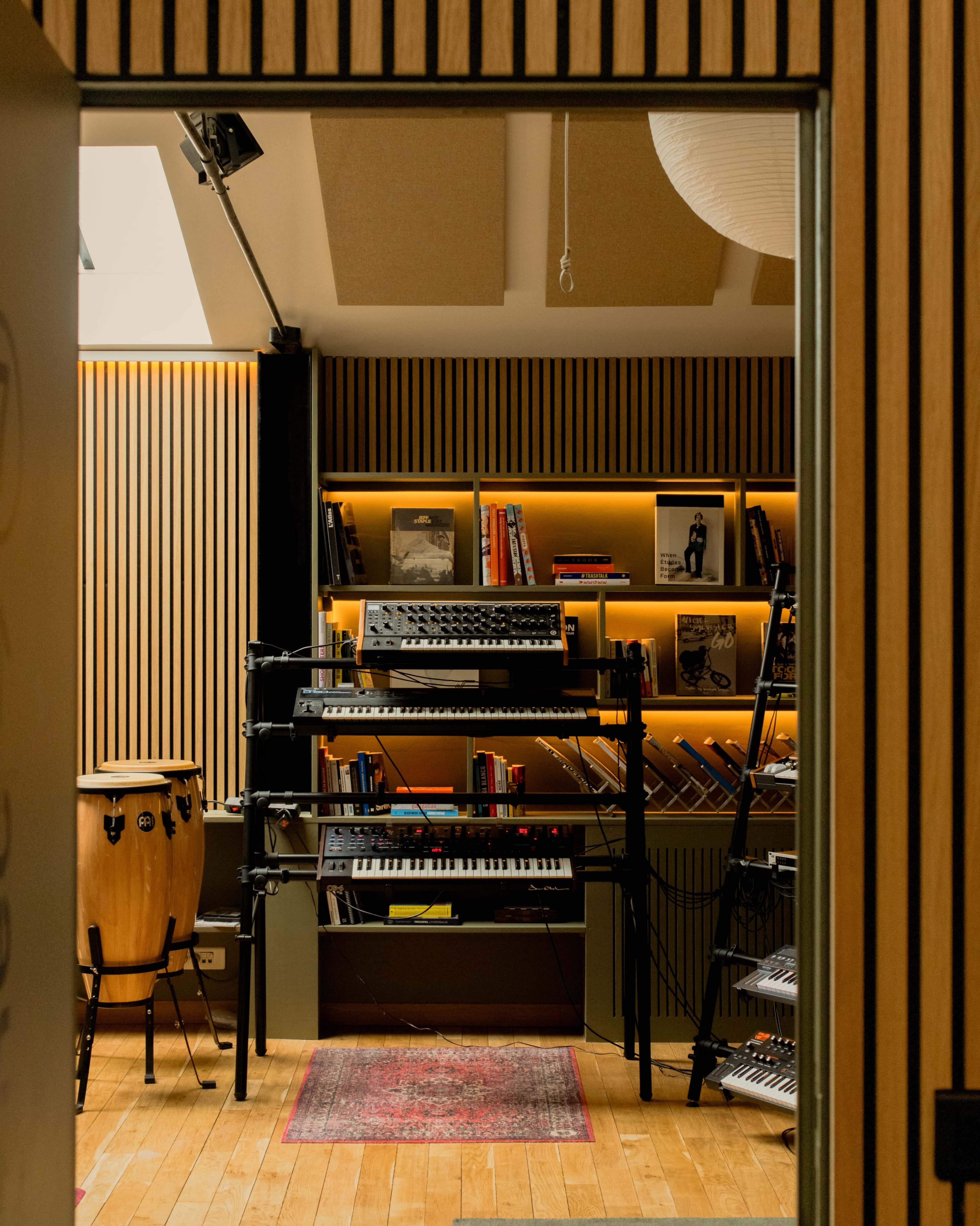 The image shows a music studio with shelves holding books and musical instruments, including keyboards and percussion, framed by wooden paneled walls and warm lighting.