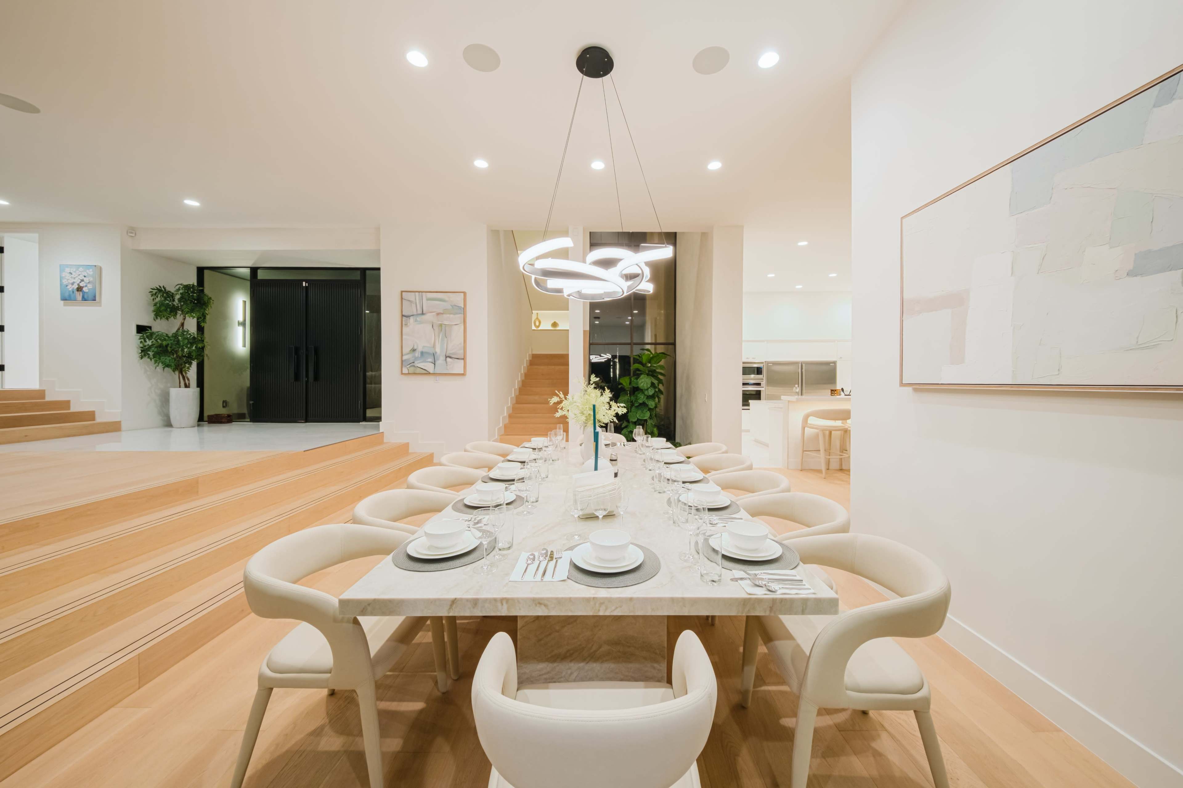 A spacious dining area features a long table set with dinnerware and a modern light fixture above, surrounded by white chairs in a well-lit room.