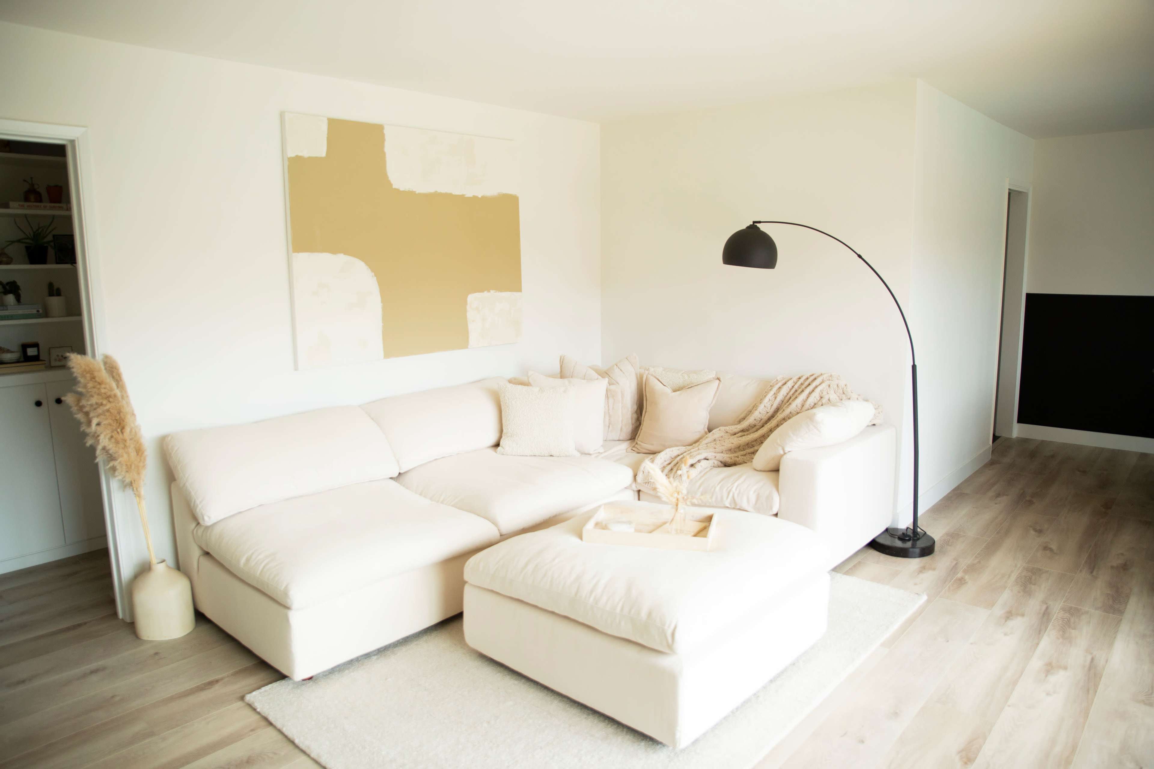 A modern living room featuring a white L-shaped sectional sofa, a floor lamp, and a beige abstract painting on the wall.
