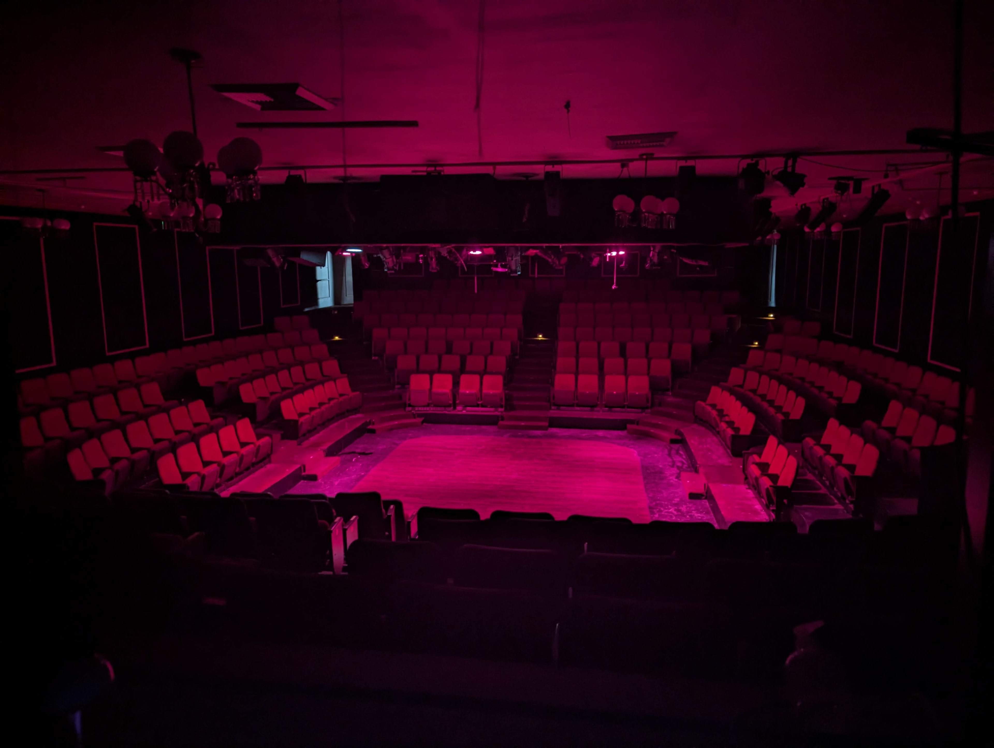 The image shows an empty theater with red seats arranged in a circular pattern around a stage, illuminated by pink lighting.