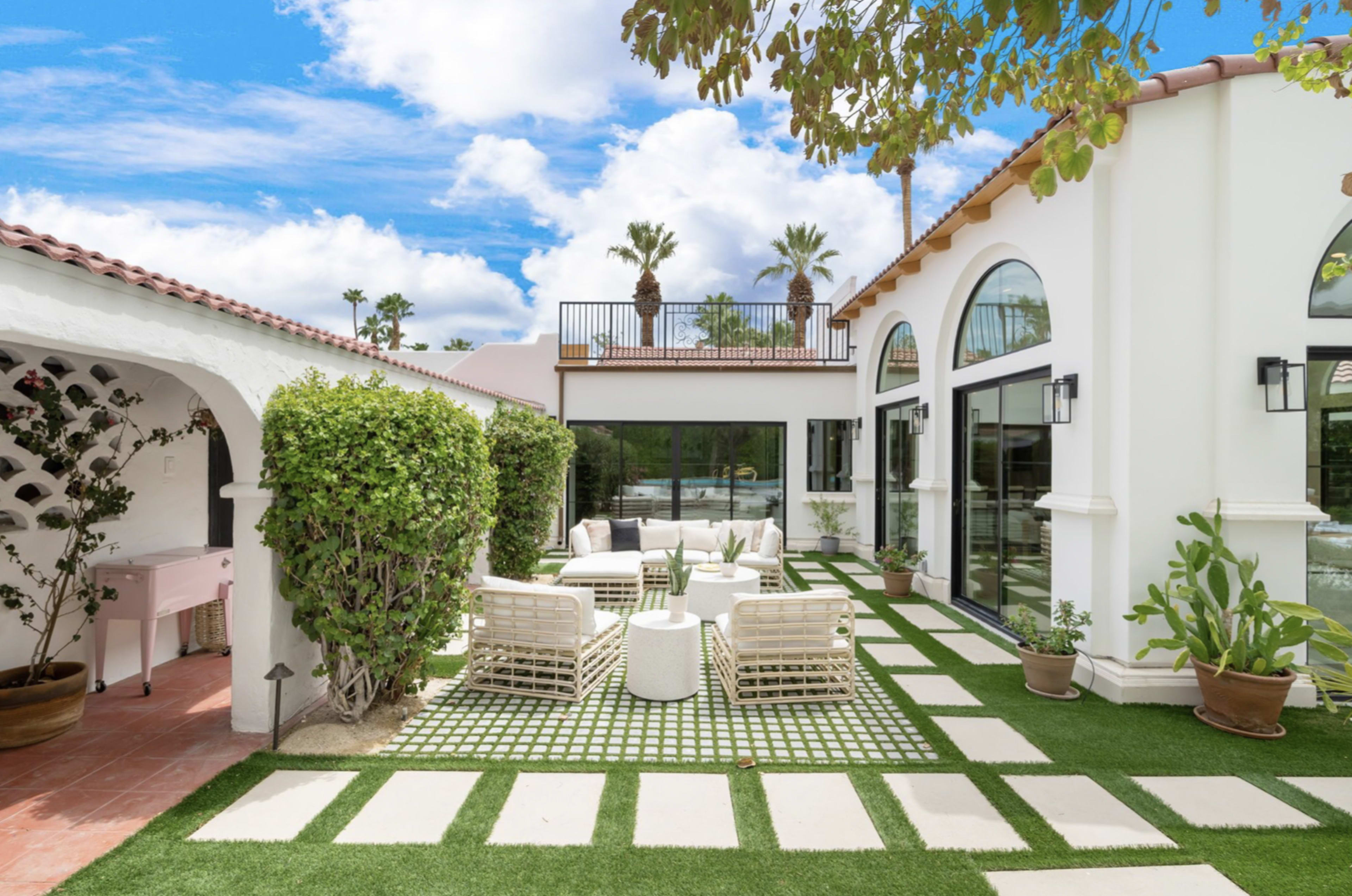 The image shows a landscaped outdoor area featuring white patio furniture, stone pathways, and lush greenery adjacent to a modern house with large windows.