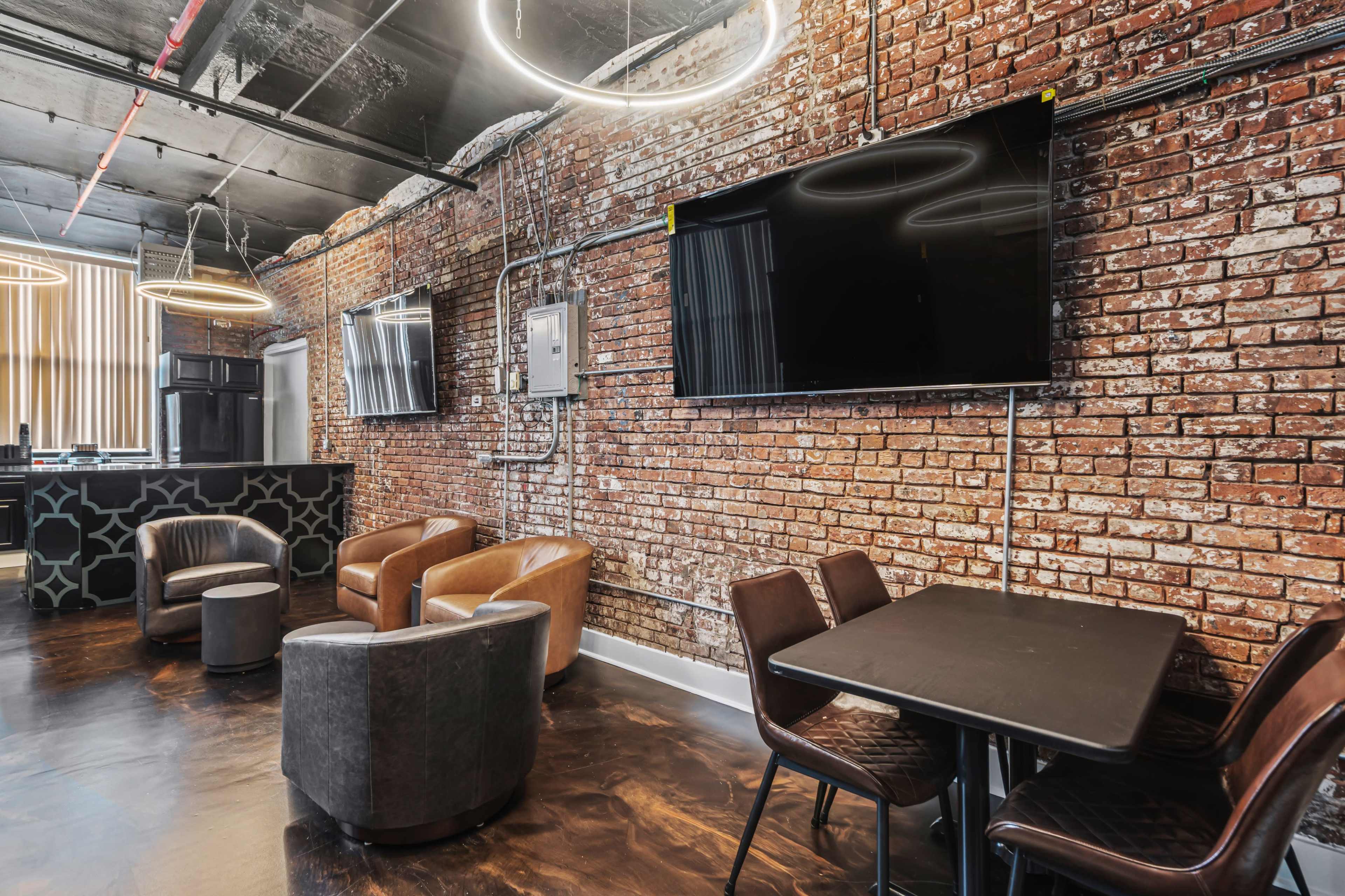 The image shows a modern lounge area with exposed brick walls, two flat-screen TVs, a bar, and seating arrangements that include brown leather chairs and a small table.