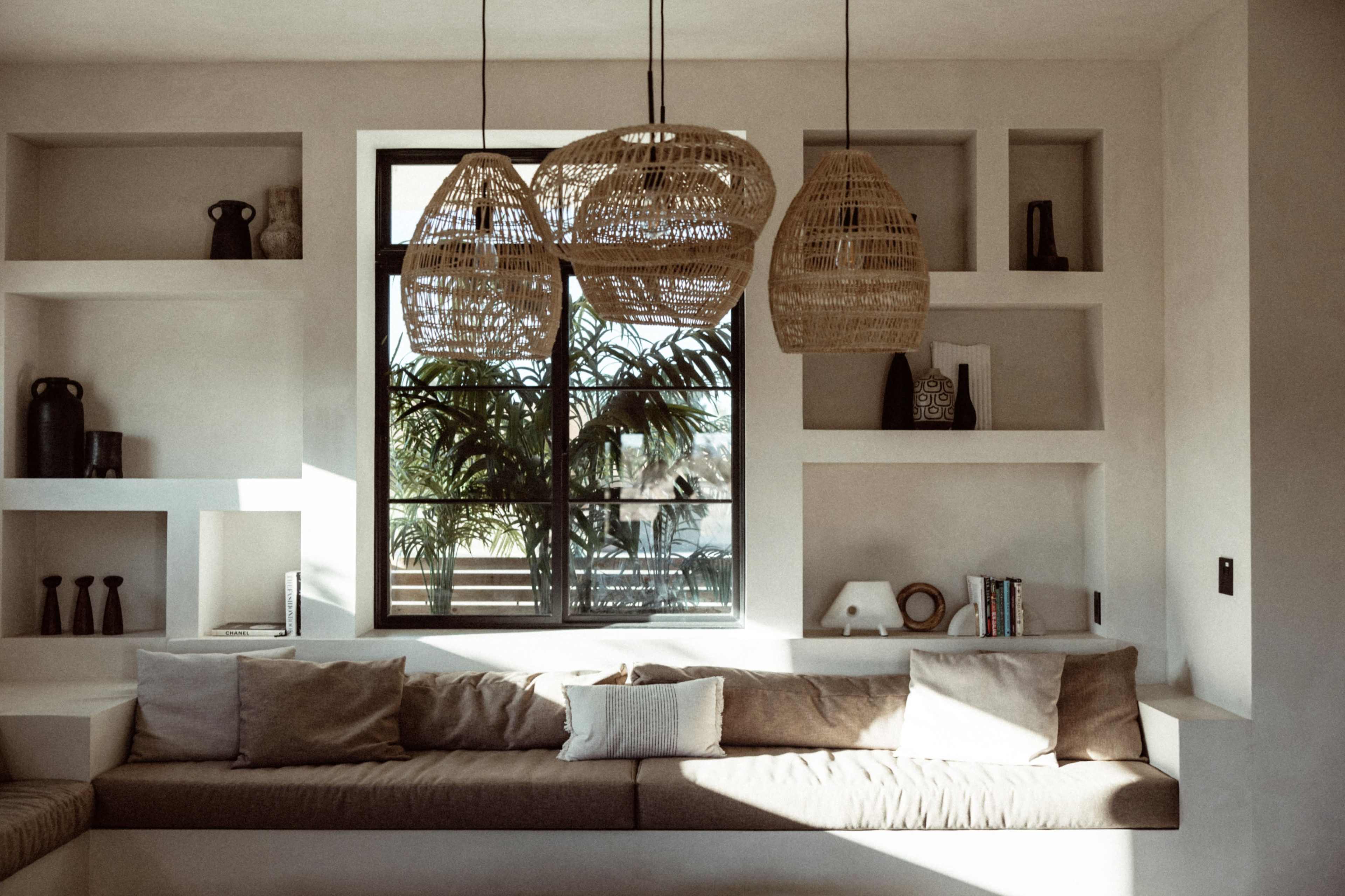 A modern interior features a cozy couch beneath three woven pendant lights, with built-in shelves displaying various decorative items and a large window revealing greenery outside.