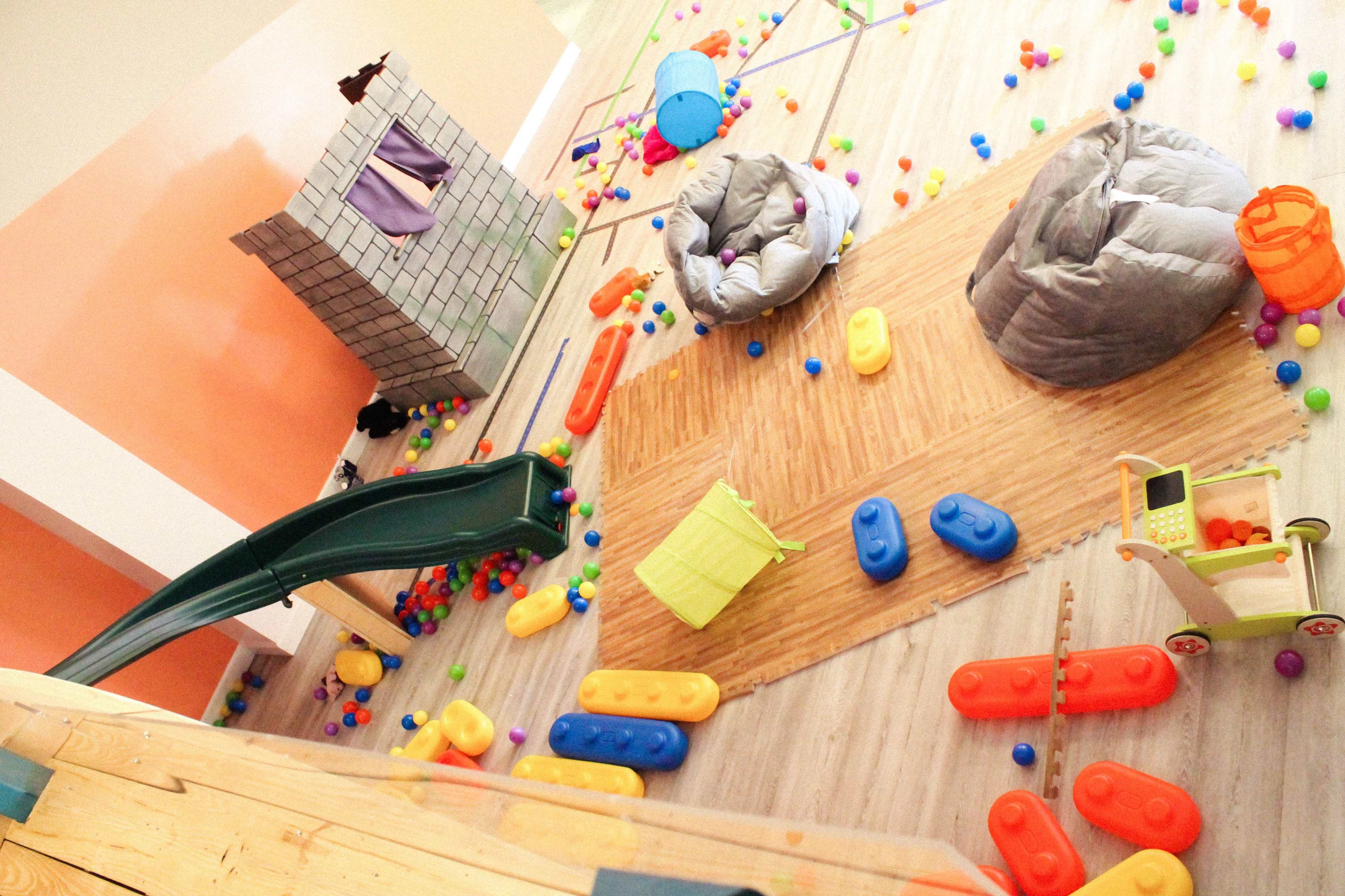 A colorful playroom is filled with play equipment, soft toys, and scattered balls across the floor.