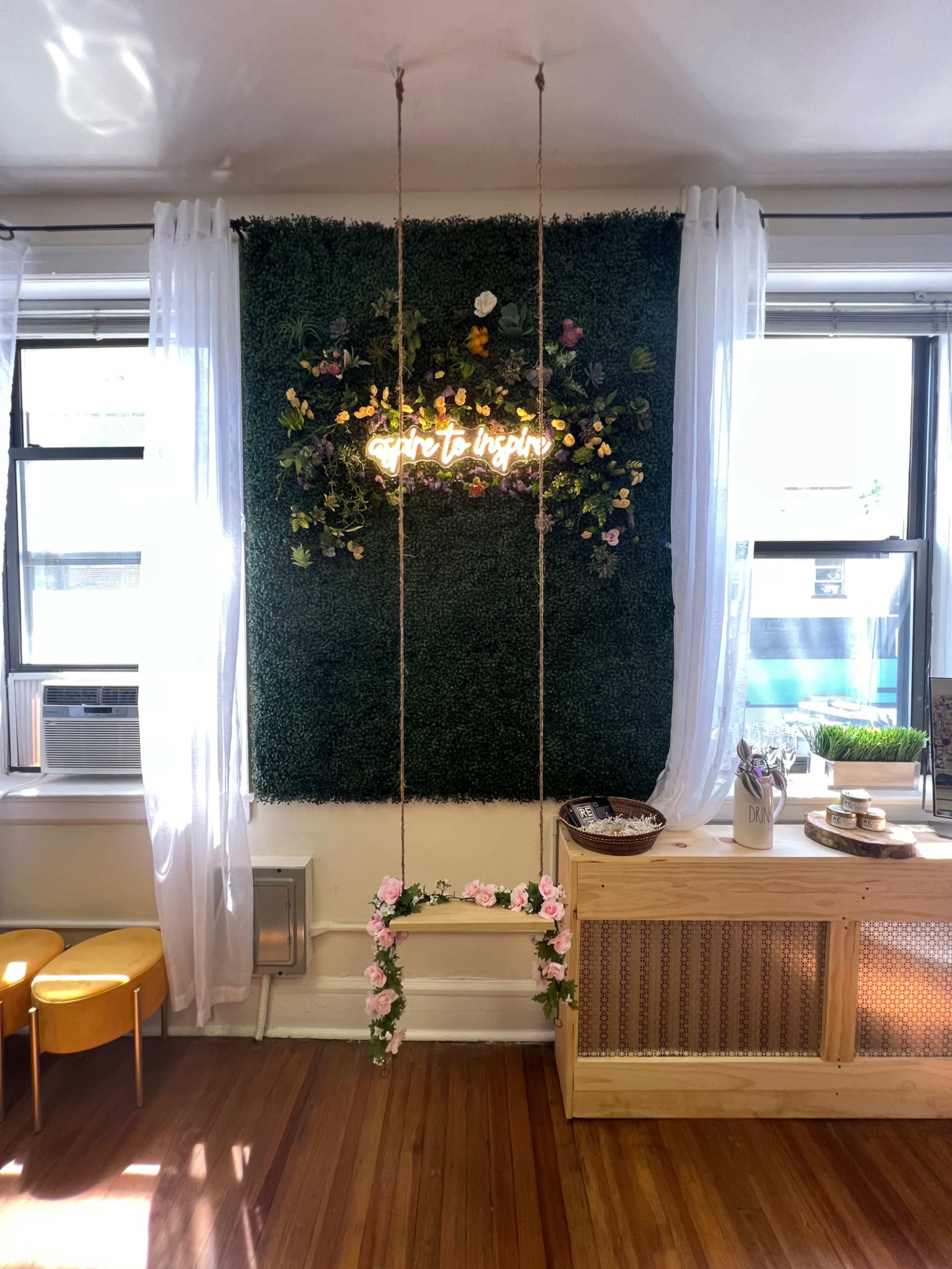 A small indoor space with a wall of greenery adorned with flowers and a neon sign reading "aspire to inspire," alongside a wooden counter and two yellow chairs.