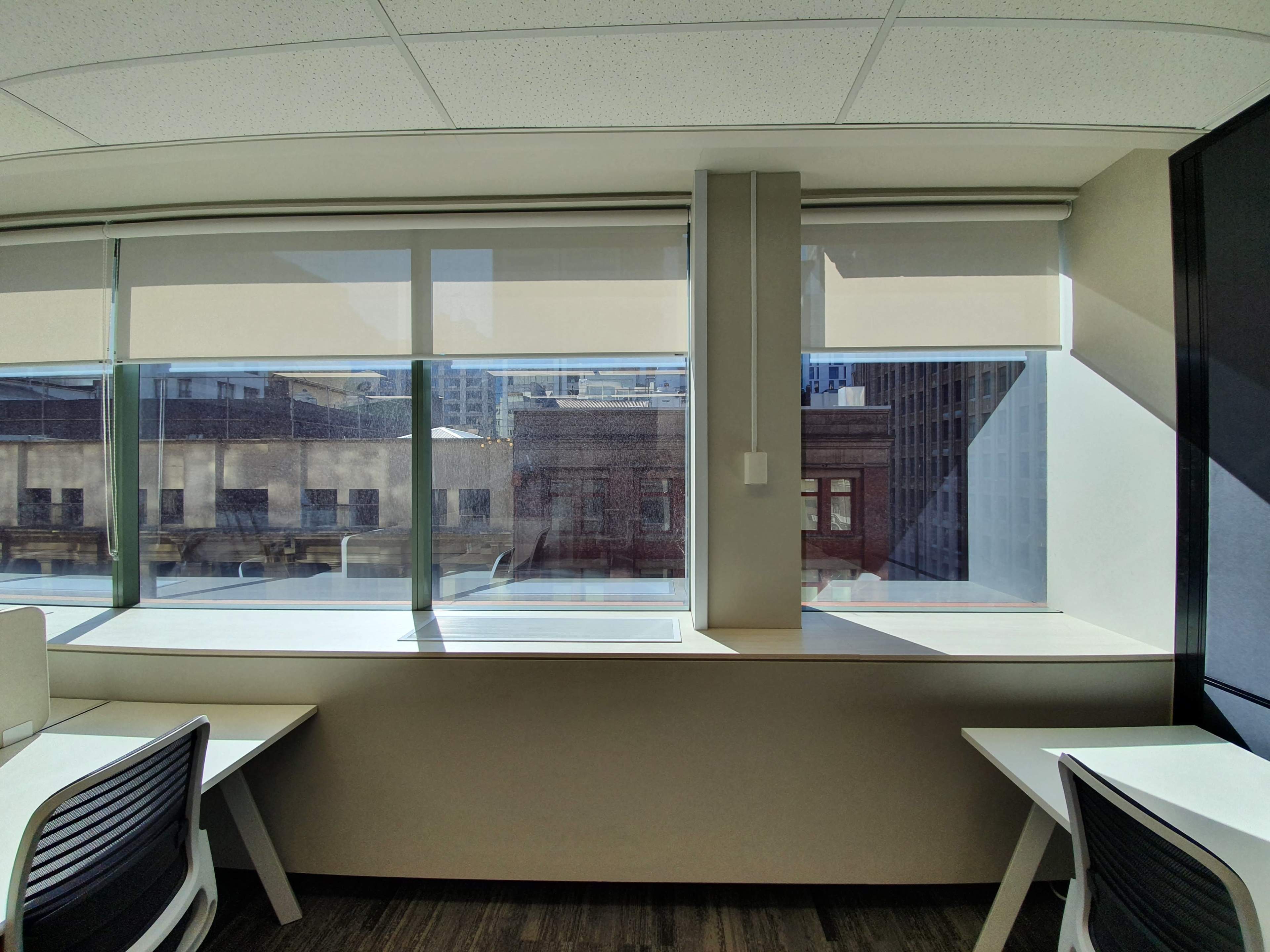 The image shows an office space with two desks positioned next to large windows, allowing natural light to illuminate the room and offering a view of a cityscape outside.