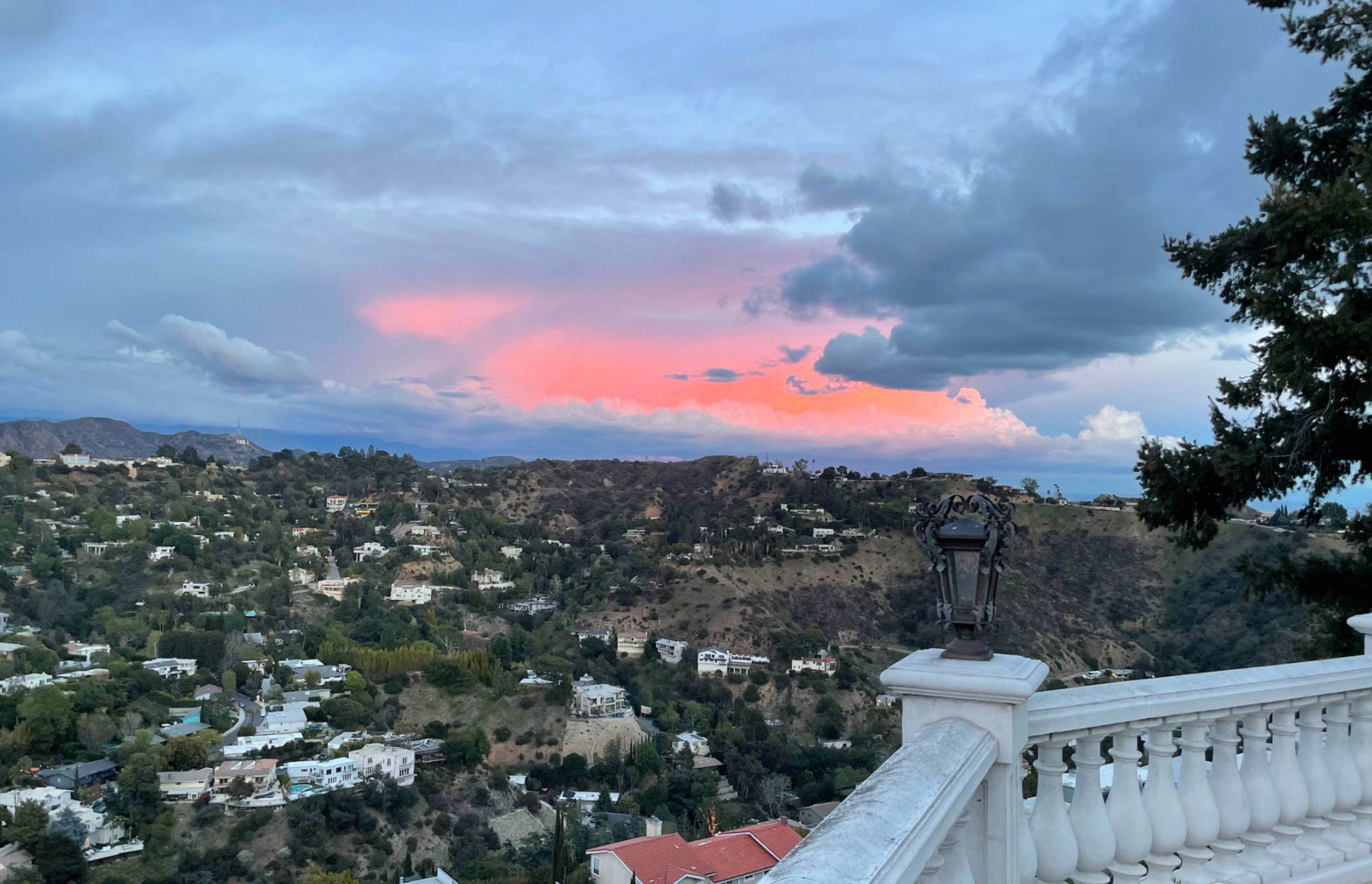 The view from a balcony shows a hilly landscape with houses beneath a colorful sky at sunset.