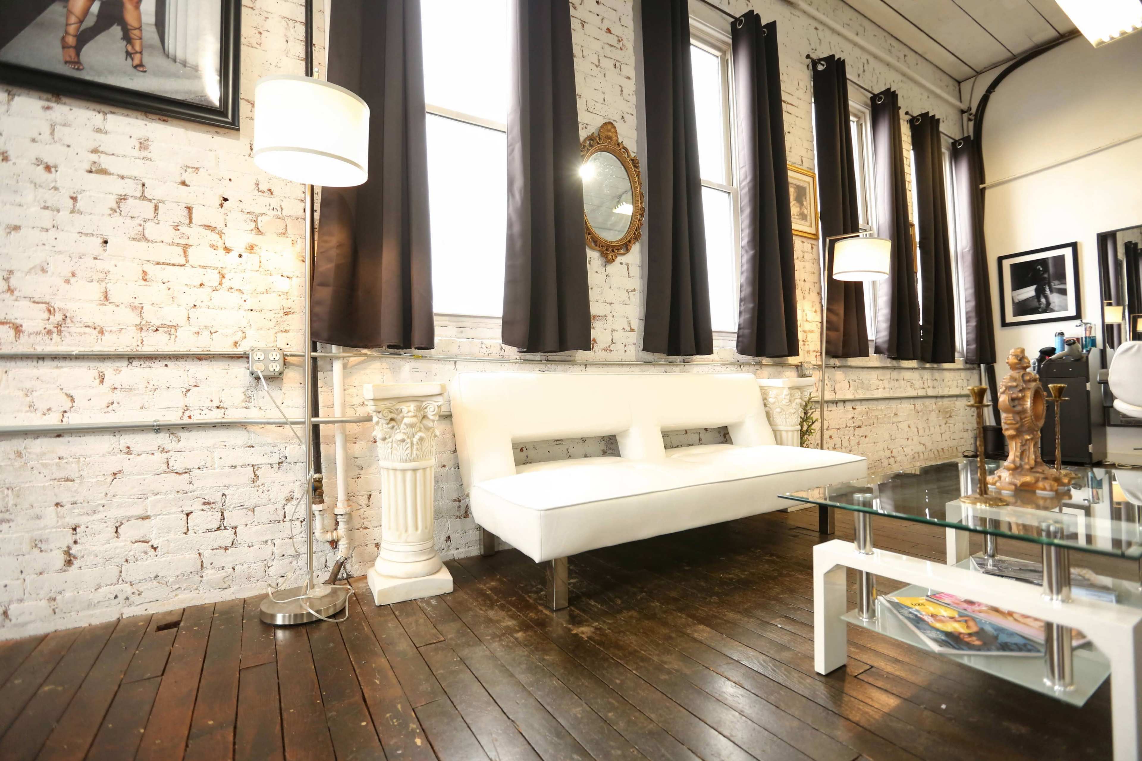 A bright room with white brick walls, a sleek white sofa, a glass coffee table, and tall windows covered by dark curtains.