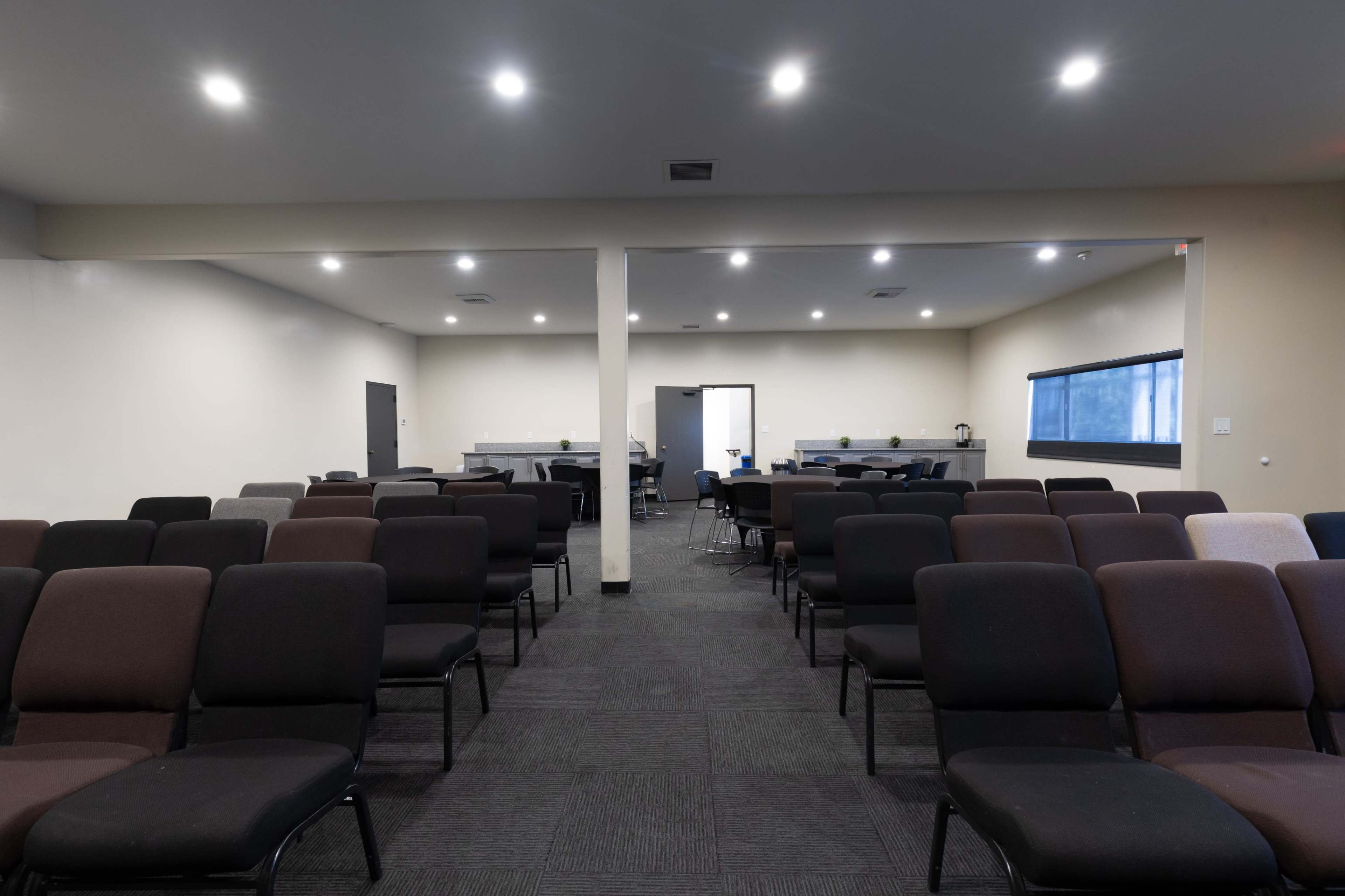 The image shows an empty conference room with rows of brown and black chairs arranged in front of a wall where there is a window and a table set up in the background.