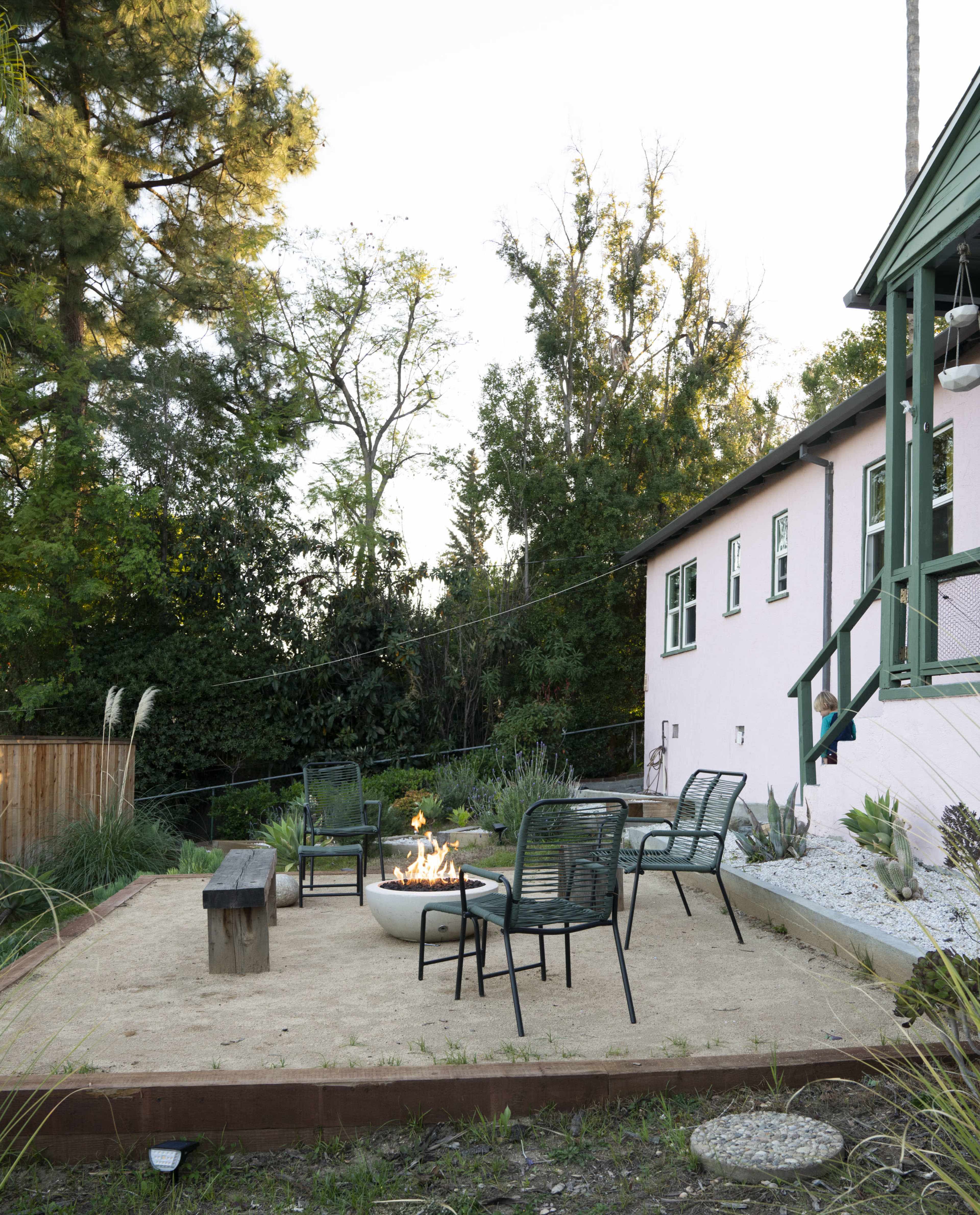 A fire pit surrounded by four chairs is situated on a sandy patch in a backyard near a light pink house.