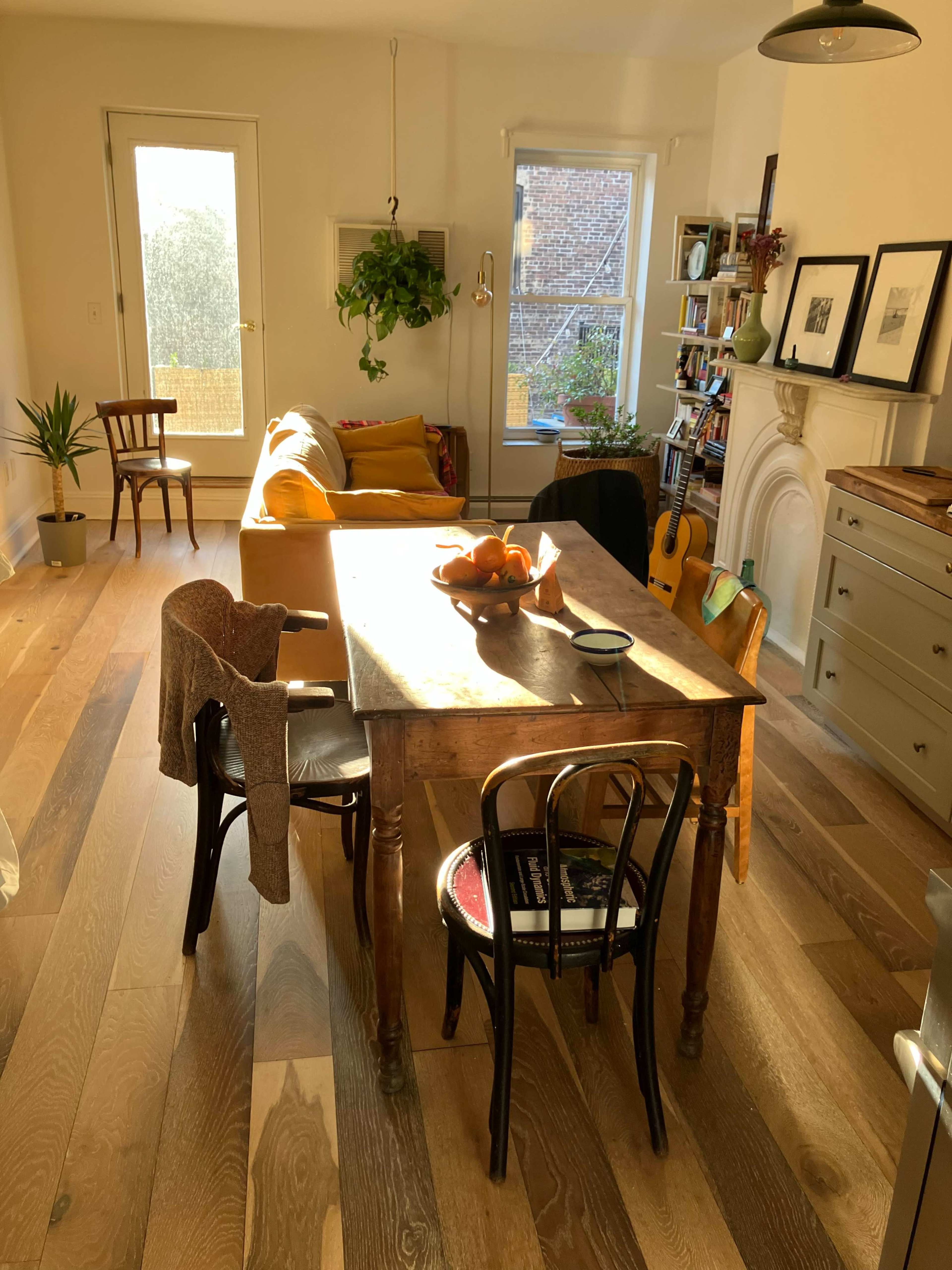 A wooden dining table is placed in a bright room with a sofa, plants, and bookshelves in the background.