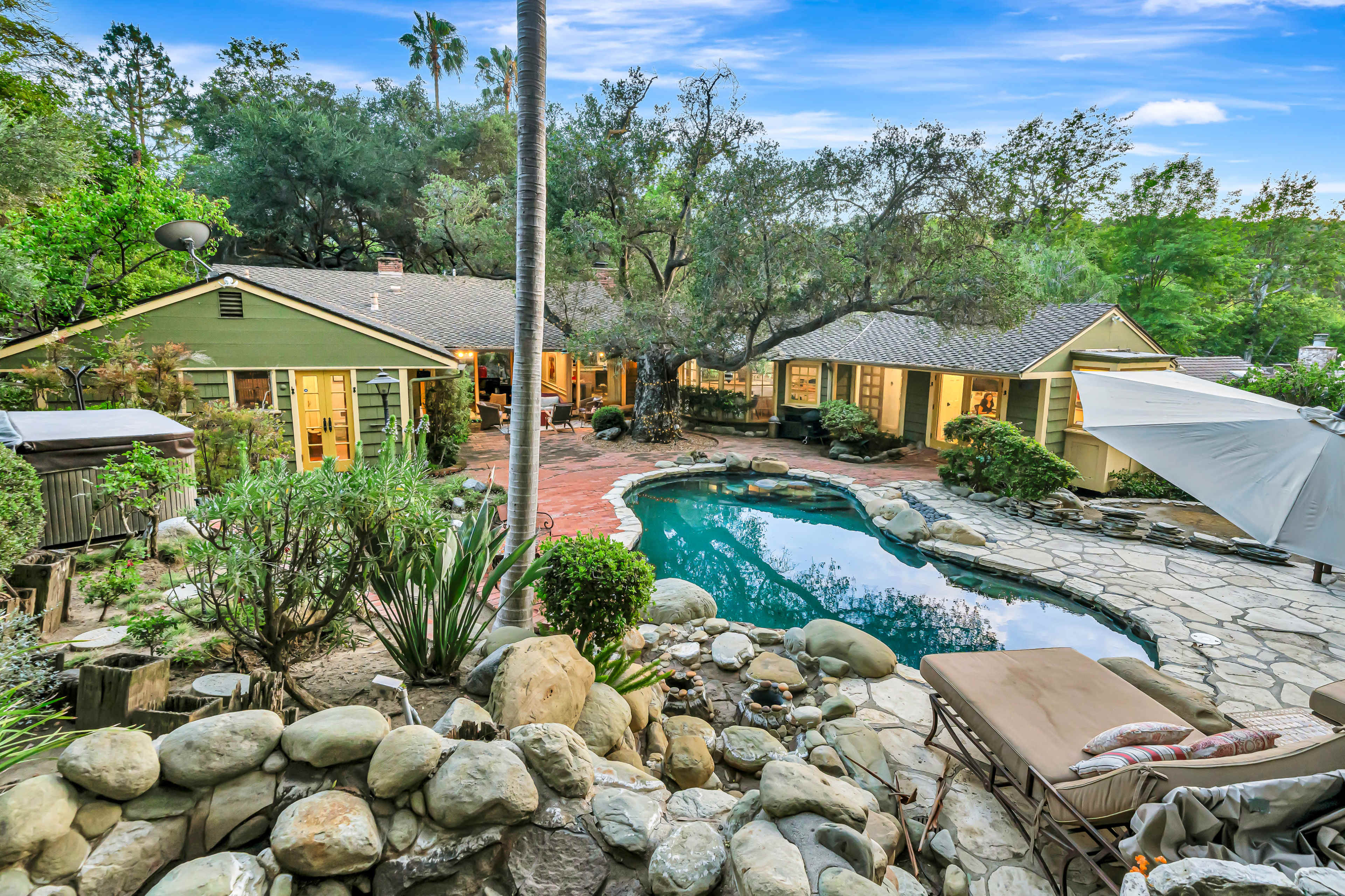 The image shows a residential property featuring a large stone-paved patio with a swimming pool, surrounded by lush greenery and multiple single-story houses.
