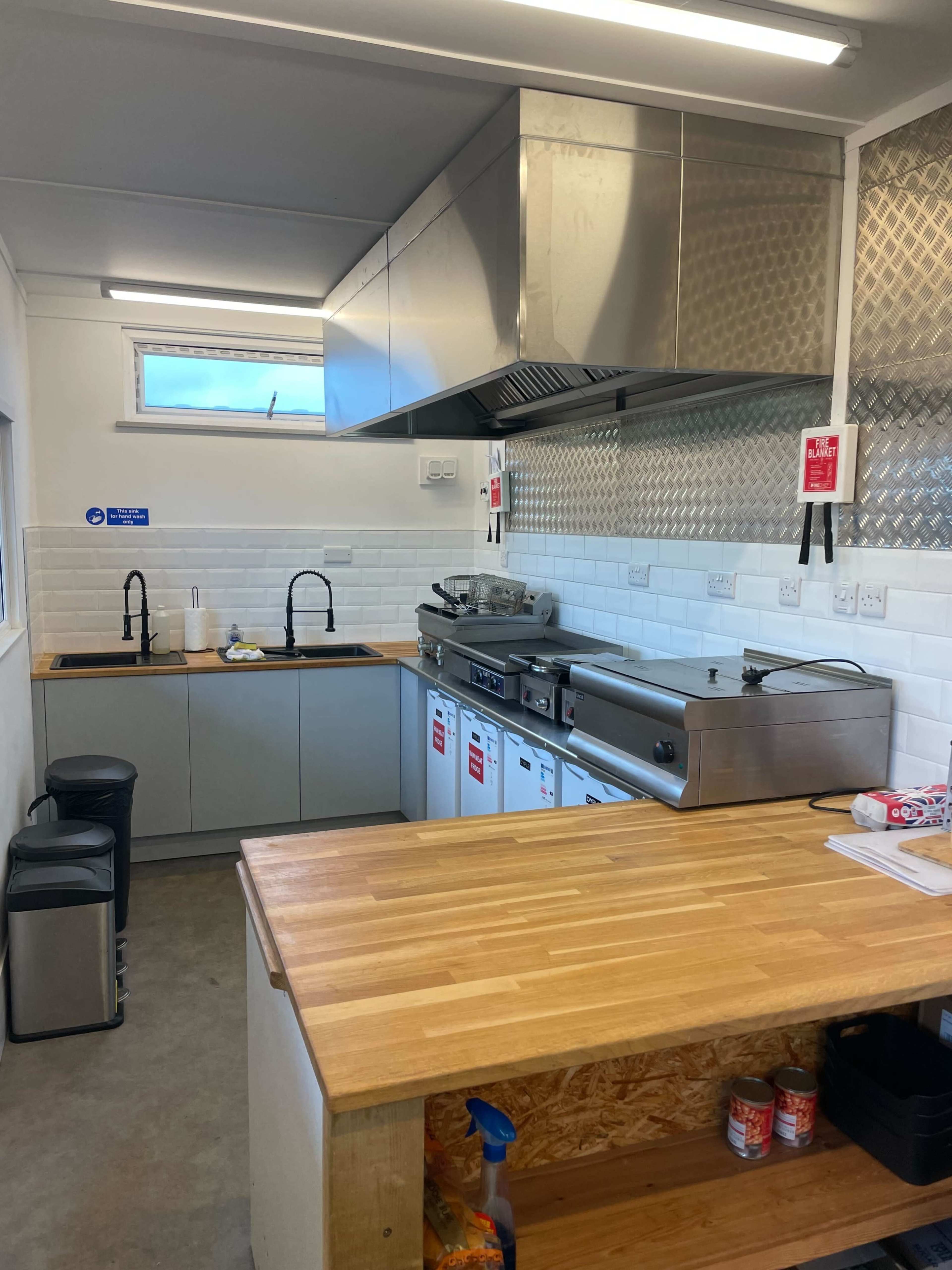 The image shows a modern kitchen with stainless steel appliances, a wooden countertop, and two sinks, along with storage cabinets and a poster on the wall.