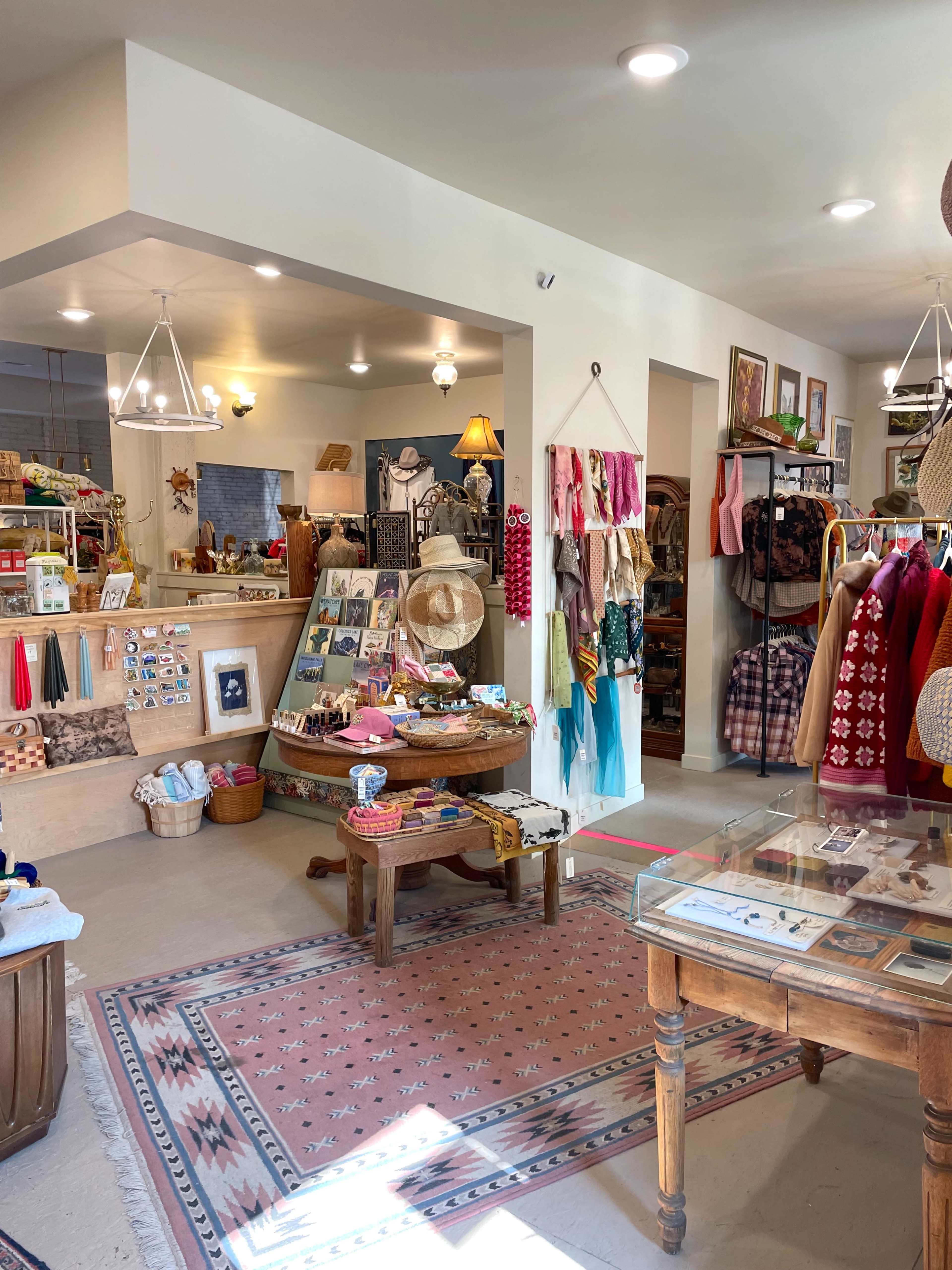 The image shows a quaint retail space filled with various handmade goods, clothing, and decorative items, with bright lighting and a warm atmosphere.