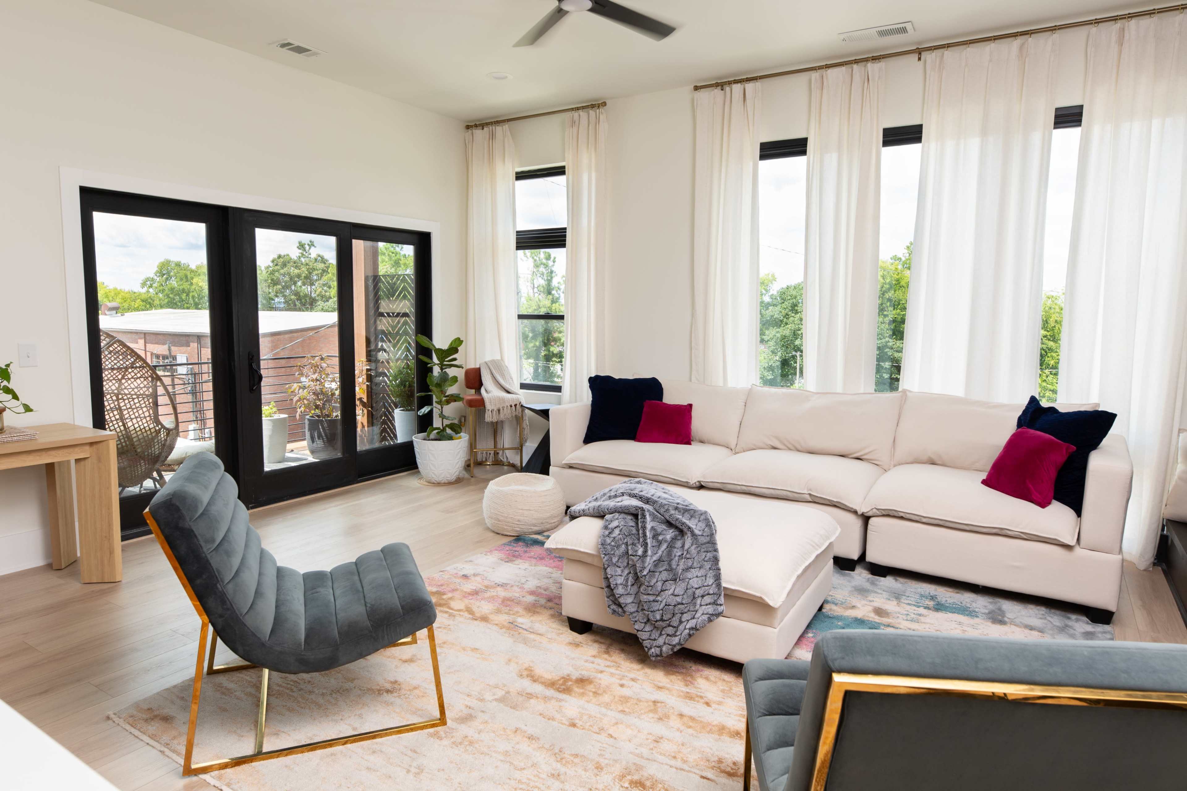 The living room features a light-colored sectional sofa with accent pillows, a cozy armchair, and large windows with sheer curtains that let in natural light.