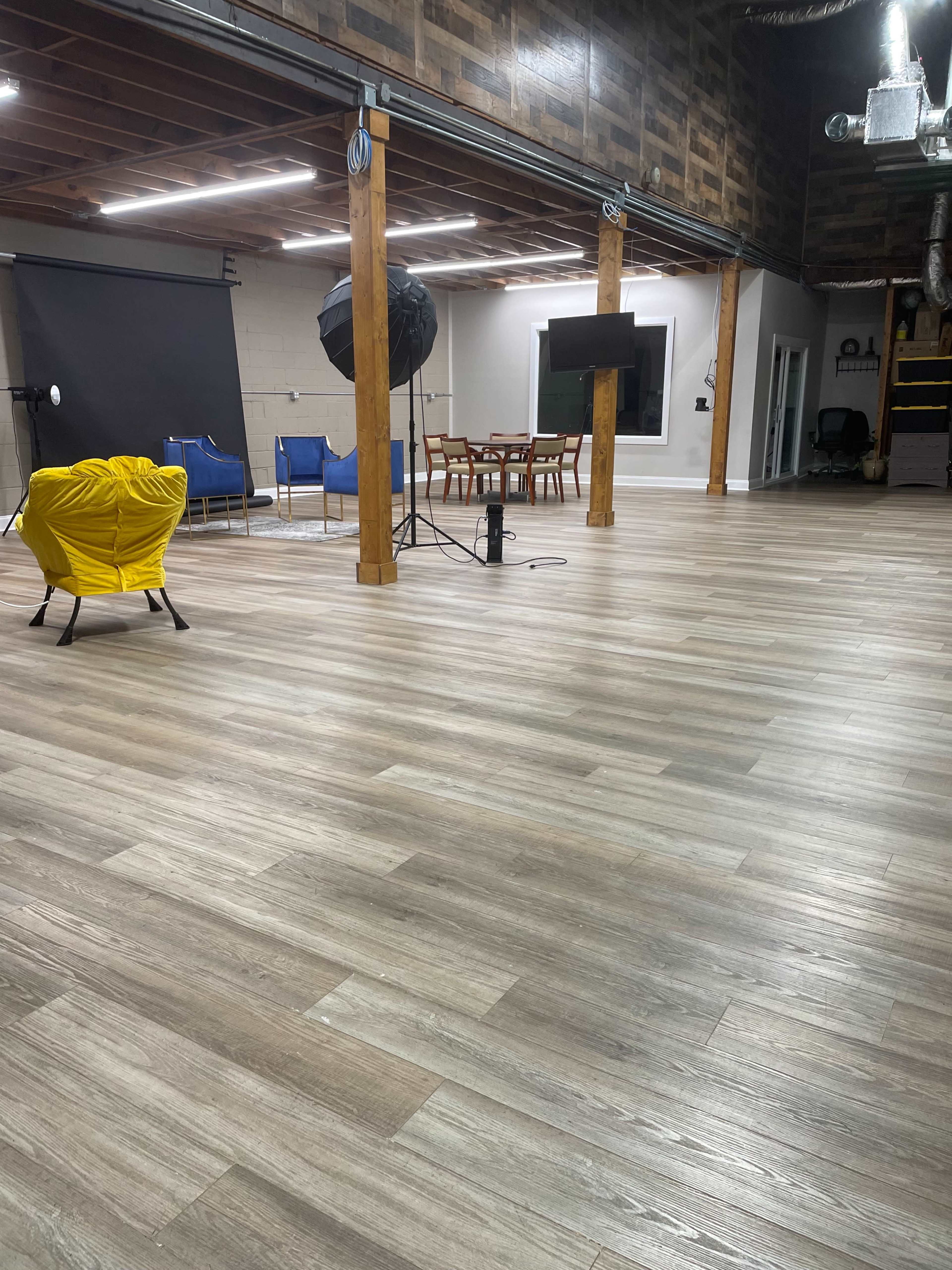 The image shows an empty studio space with wooden flooring, equipped with lighting equipment, chairs, a dining table, and a television mounted on the wall.
