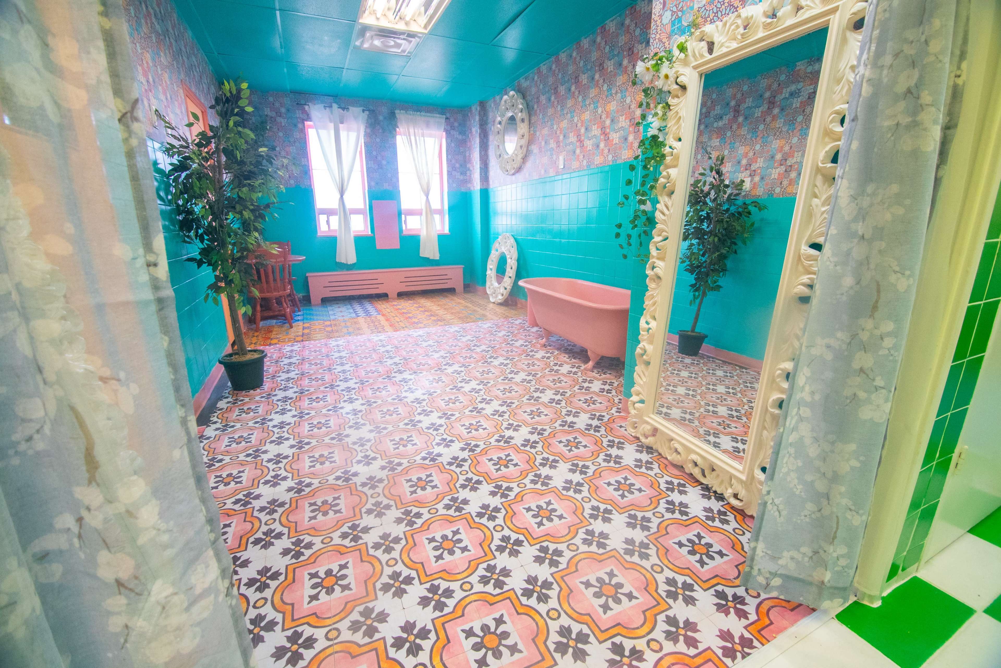 A colorful bathroom with patterned tiles, a pink bathtub, a large ornate mirror, and plants arranged in the corners.