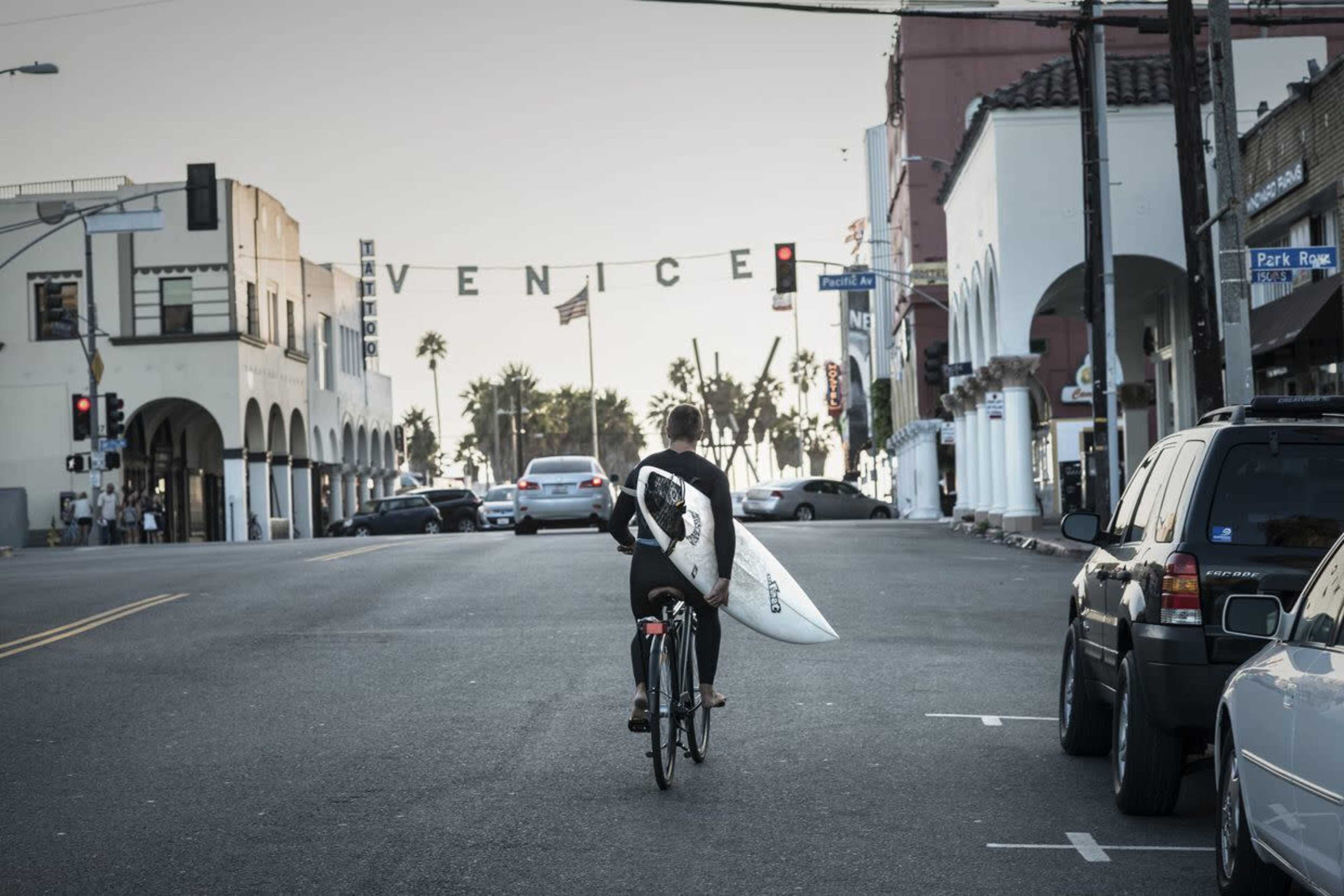 A person carrying a surfboard walks a bicycle down a street in Venice, California, with palm trees and buildings in the background.